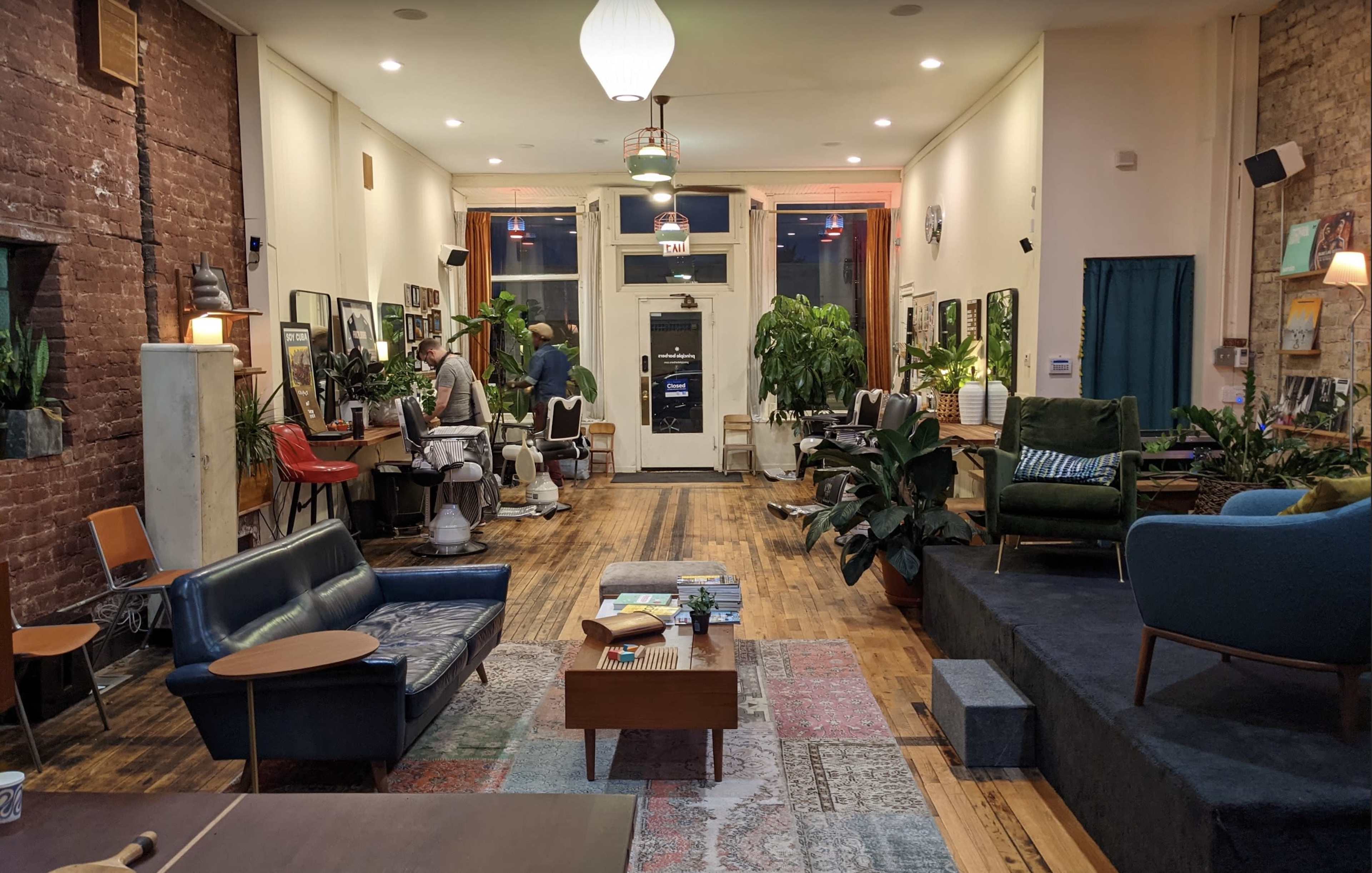 The image depicts a cozy co-working space featuring multiple seating areas, plants, and workstations with people engaged in various activities.