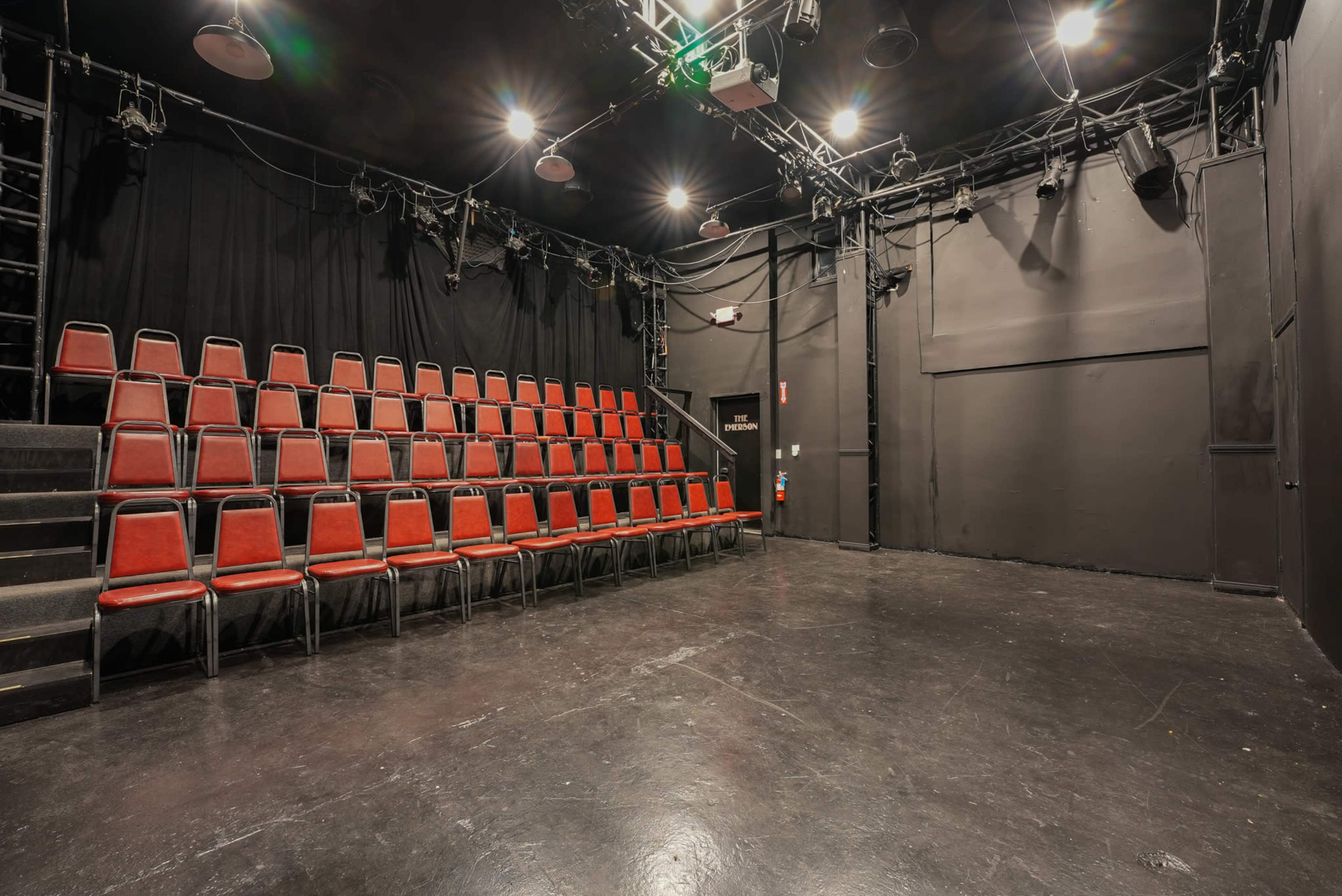 A small theater space features rows of empty red chairs against black walls and a dark floor, with stage lighting fixtures overhead.