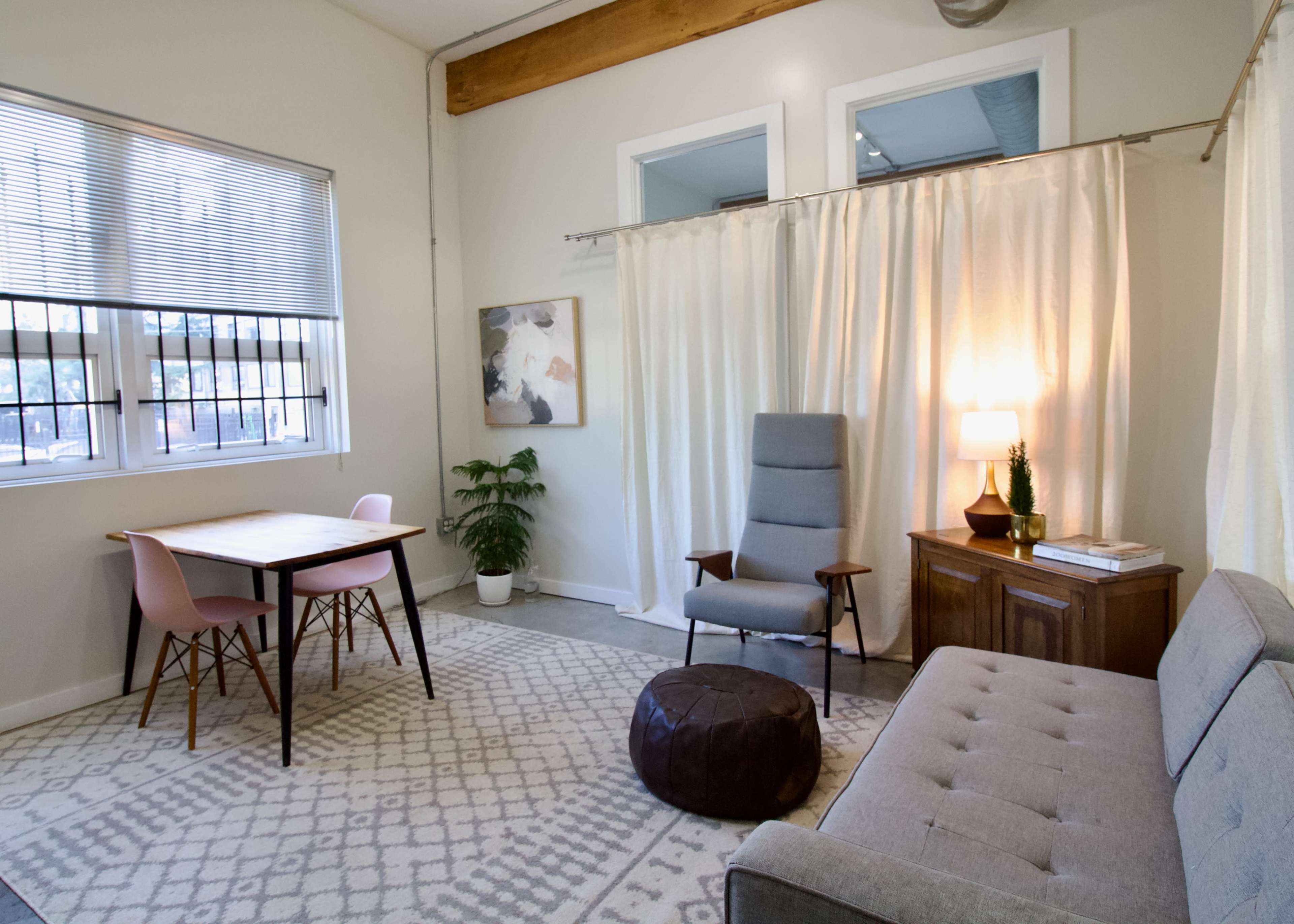 The image depicts a cozy, well-lit room featuring a gray sofa, a wooden table with chairs, a plant, and a folding curtain separating a chair and lamp setup.