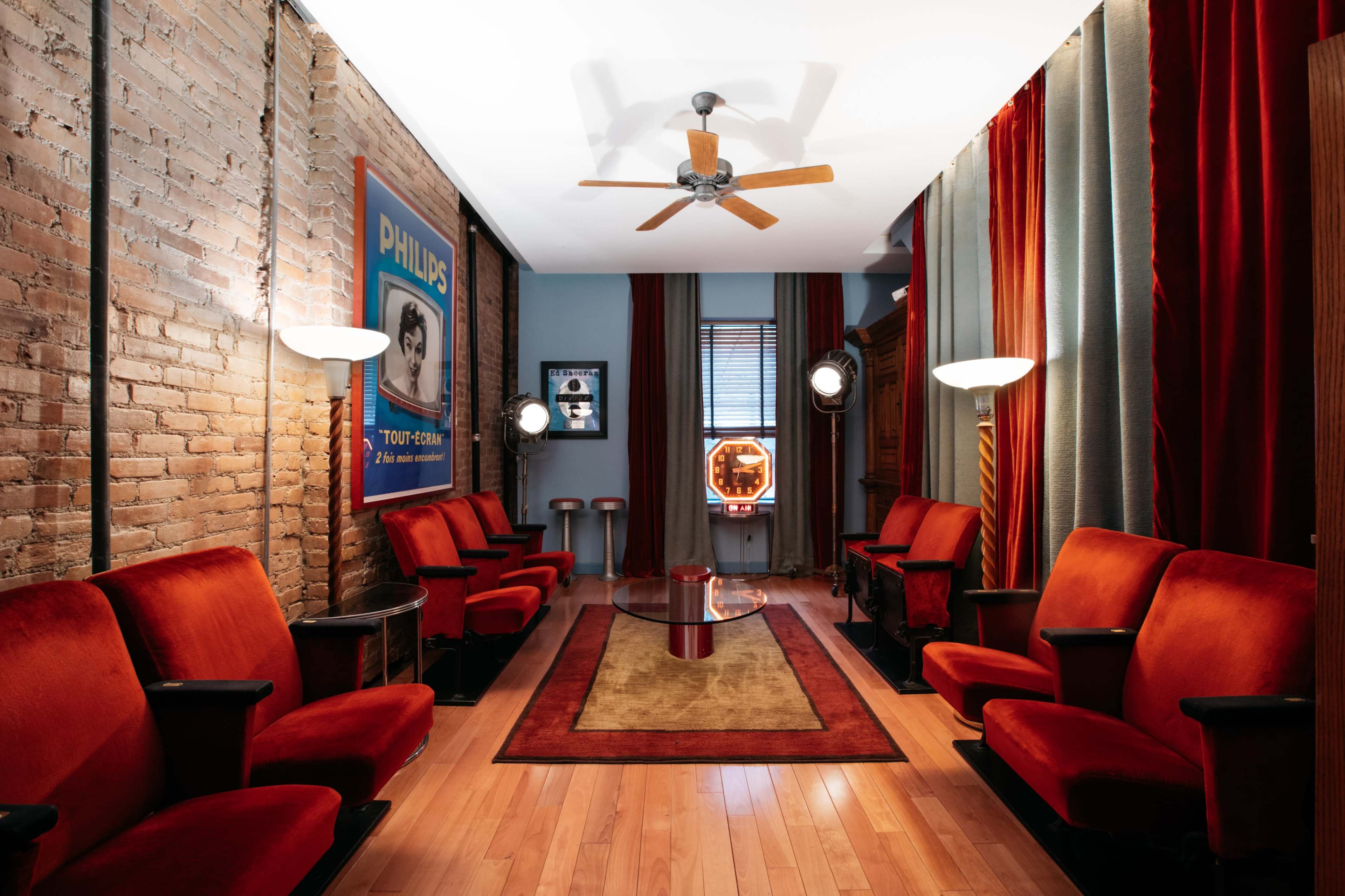 The room features vintage red velvet seating arranged along a wooden floor, with warm lighting and decorative curtains beside a framed advertisement on the wall.