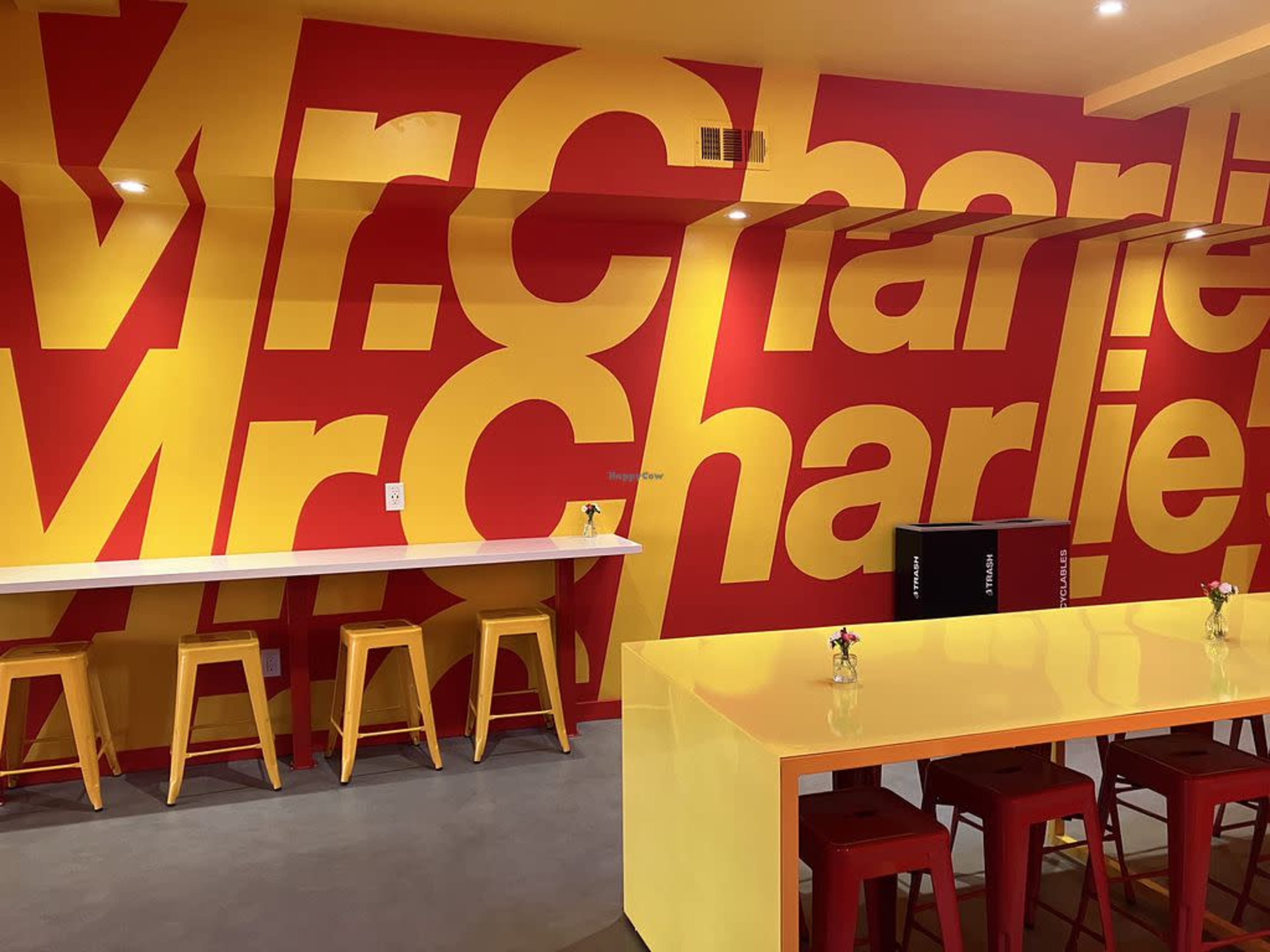 The image shows a brightly colored café with a bold red and yellow wall design featuring the name "Mr. Charlie," along with seating at a long white counter and small tables with flowers.