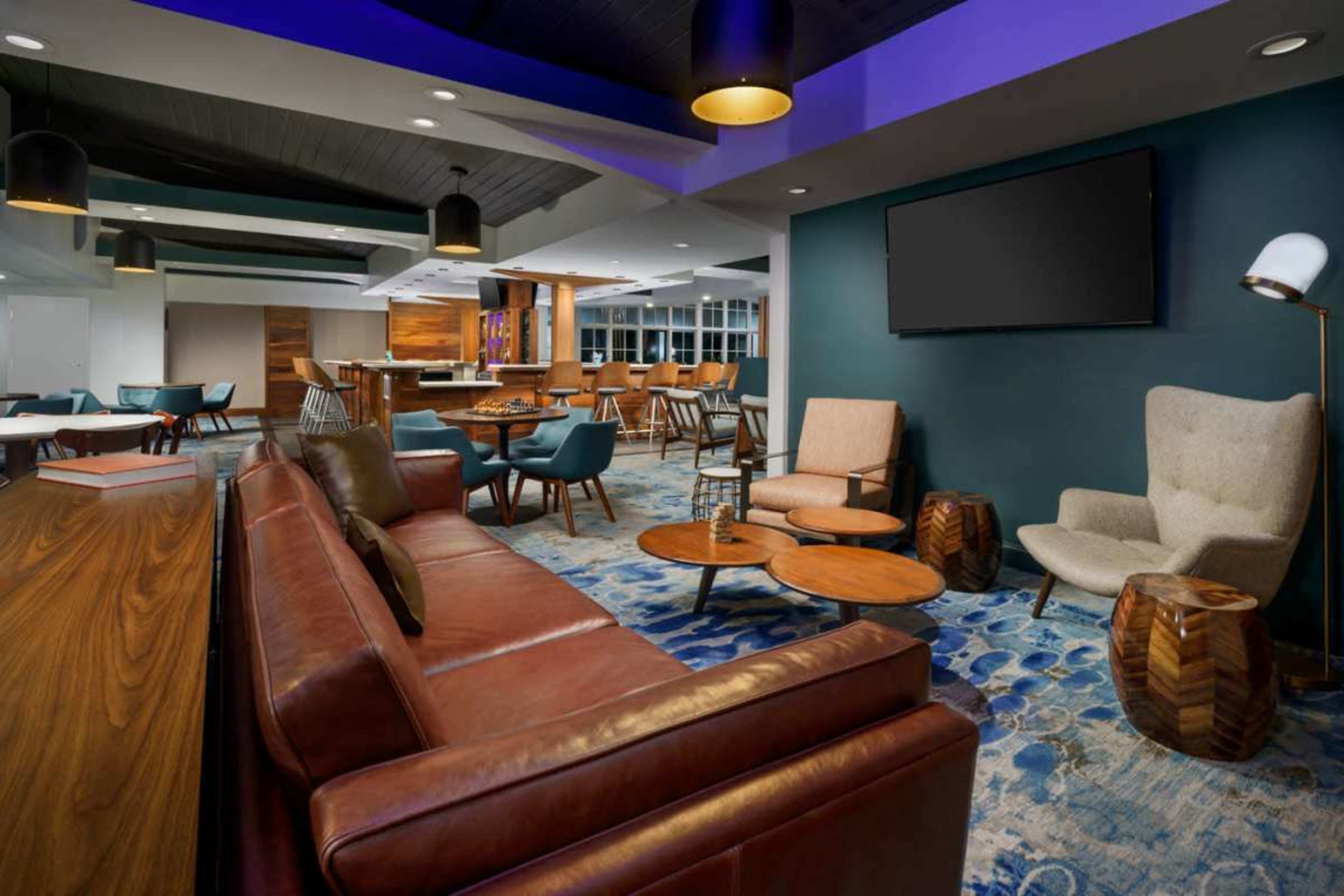 The image shows a modern lounge area with brown leather sofas, wooden tables, and various seating options, highlighted by blue and purple lighting.