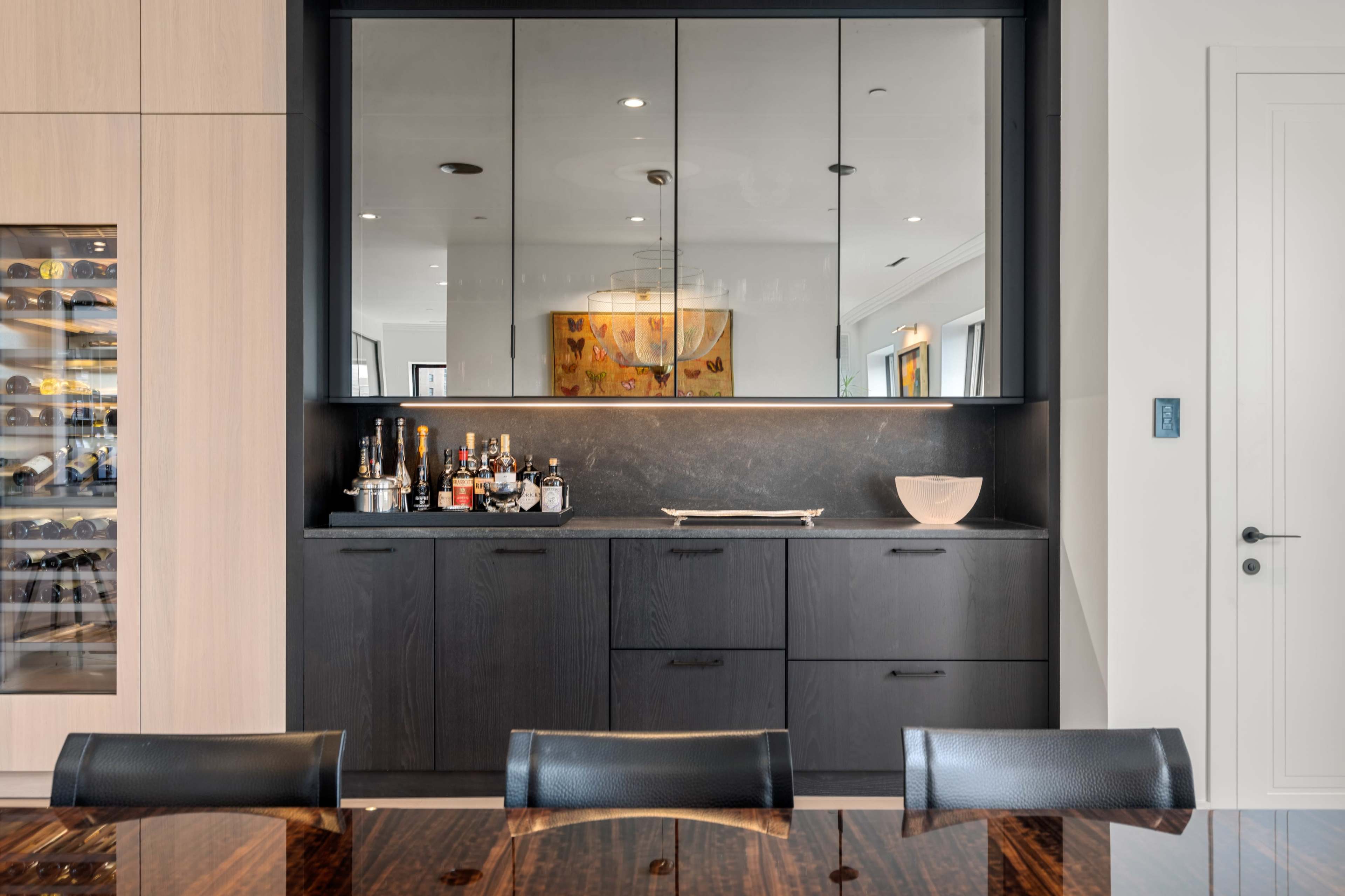 A modern kitchen bar area with a dark cabinetry unit, a mirrored backsplash, and various bottles displayed on the countertop.