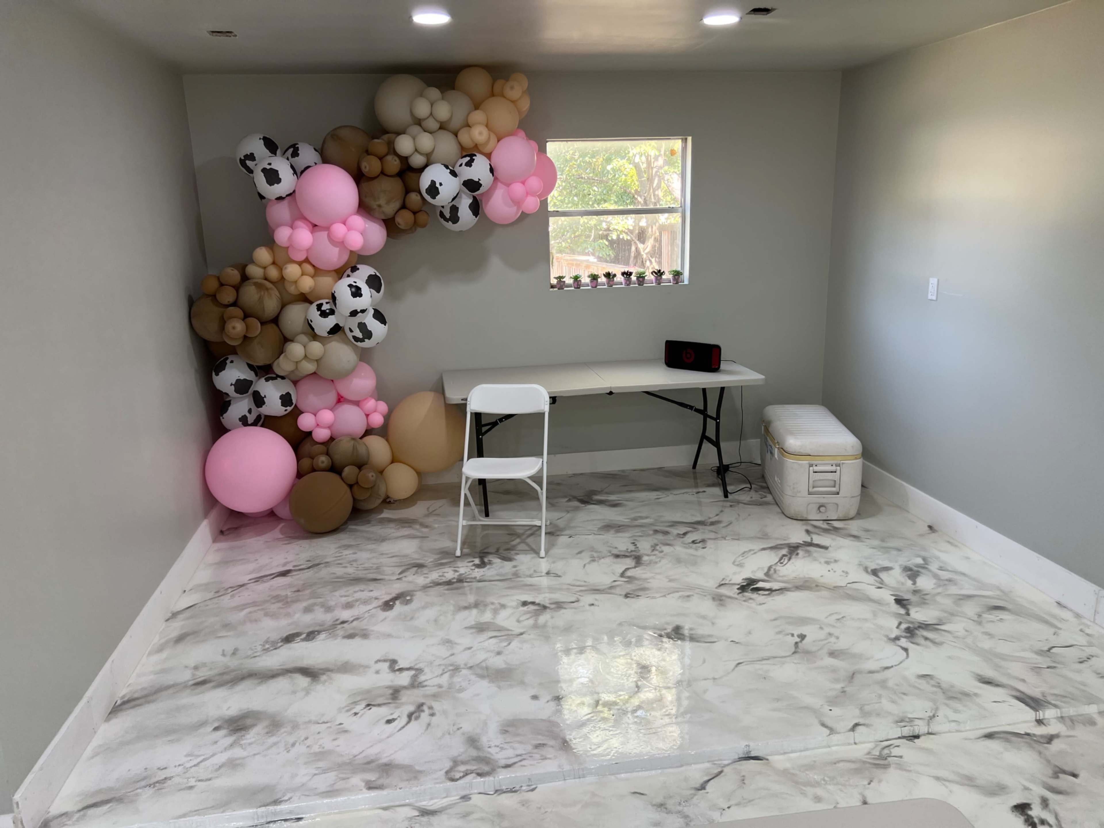 The room features a table with a white chair, a cooler, and a colorful balloon arrangement in the corner, all set against a marble-patterned floor.