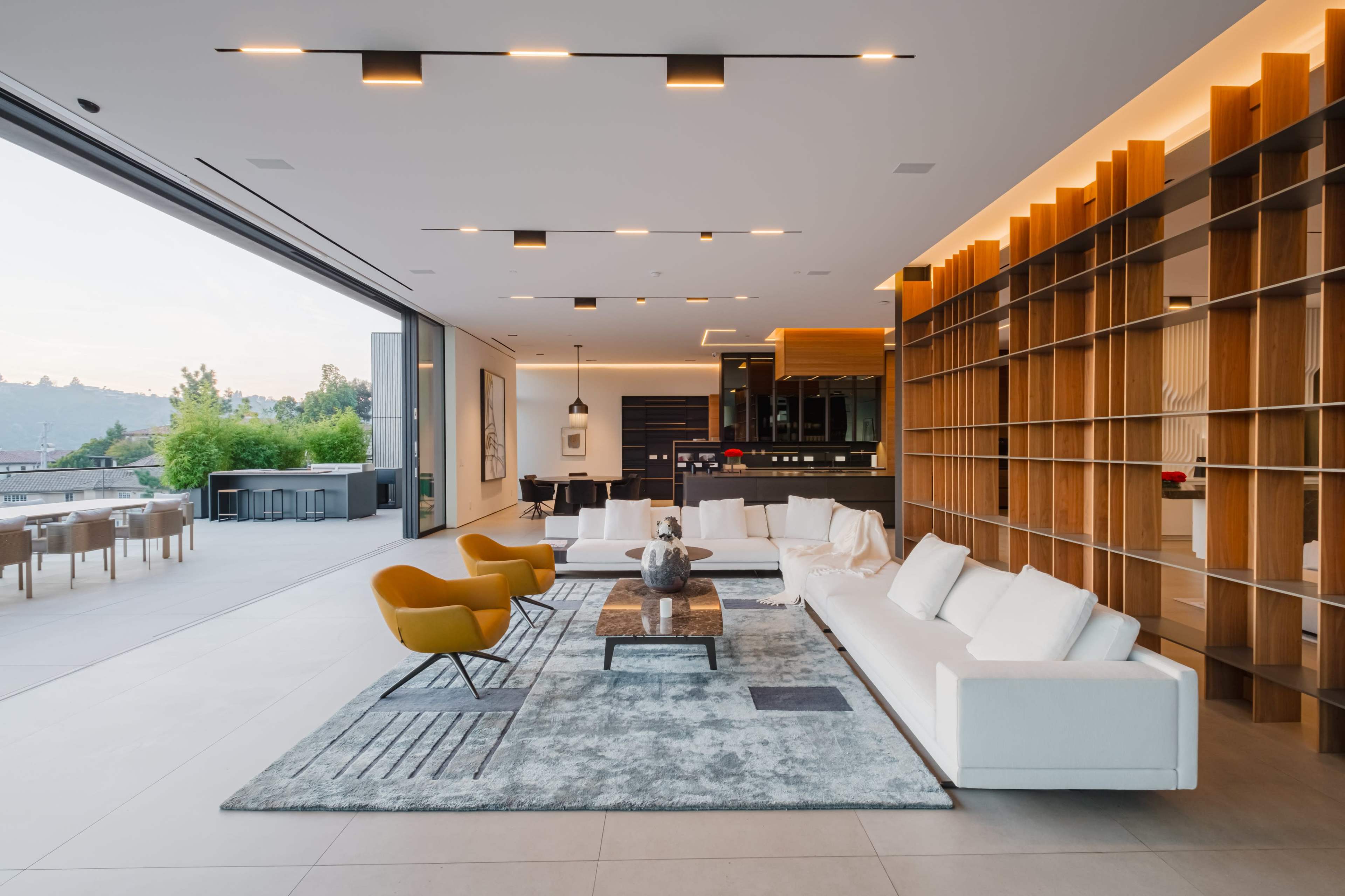 A spacious modern living room features a large white sofa, two yellow armchairs, a wooden bookshelf, and an open view of the outdoors through floor-to-ceiling windows.