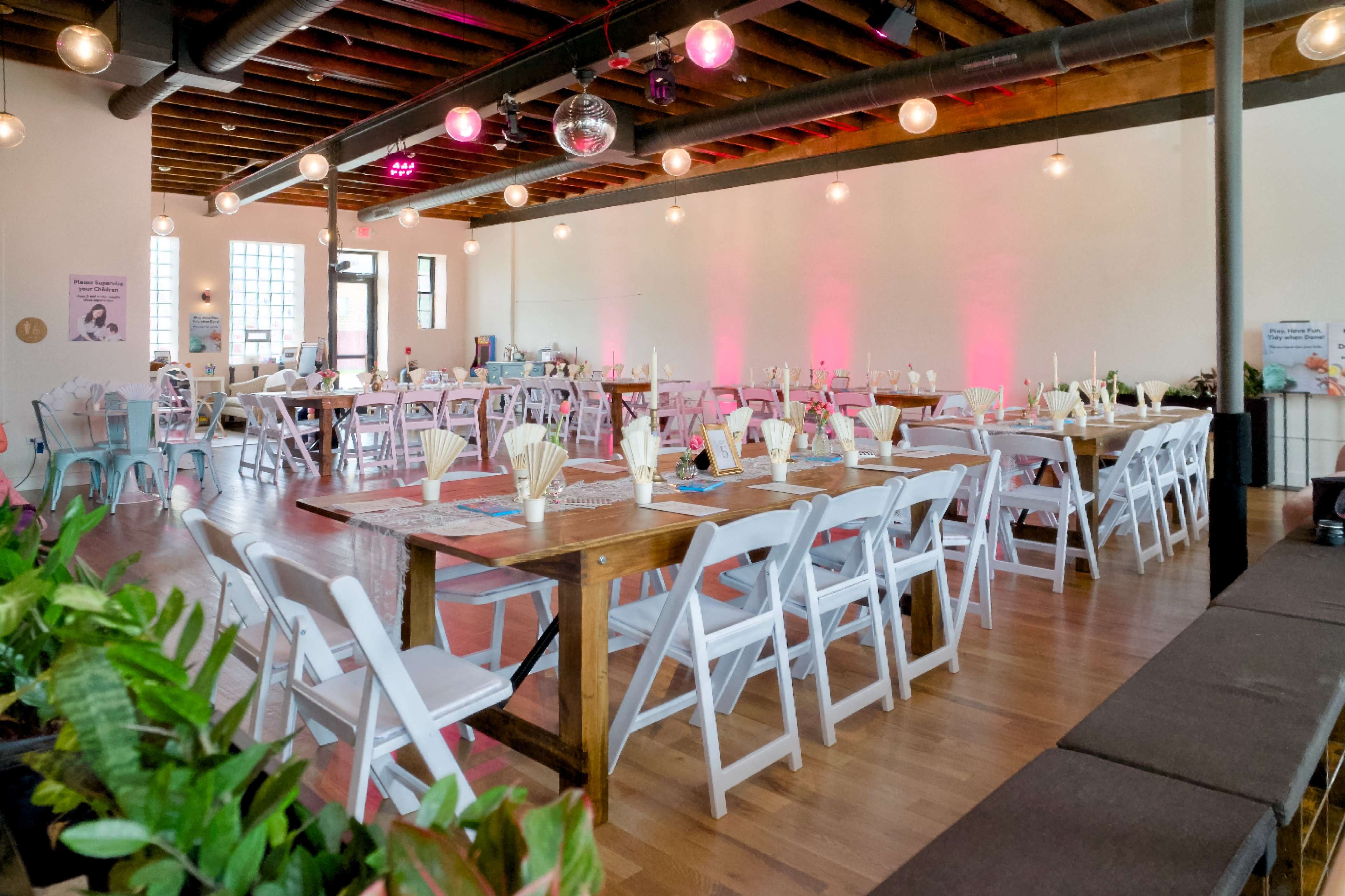 The scene shows a spacious event venue set up for a gathering, featuring rows of tables with white chairs and a mix of wooden and colorful accents, illuminated by overhead lights.