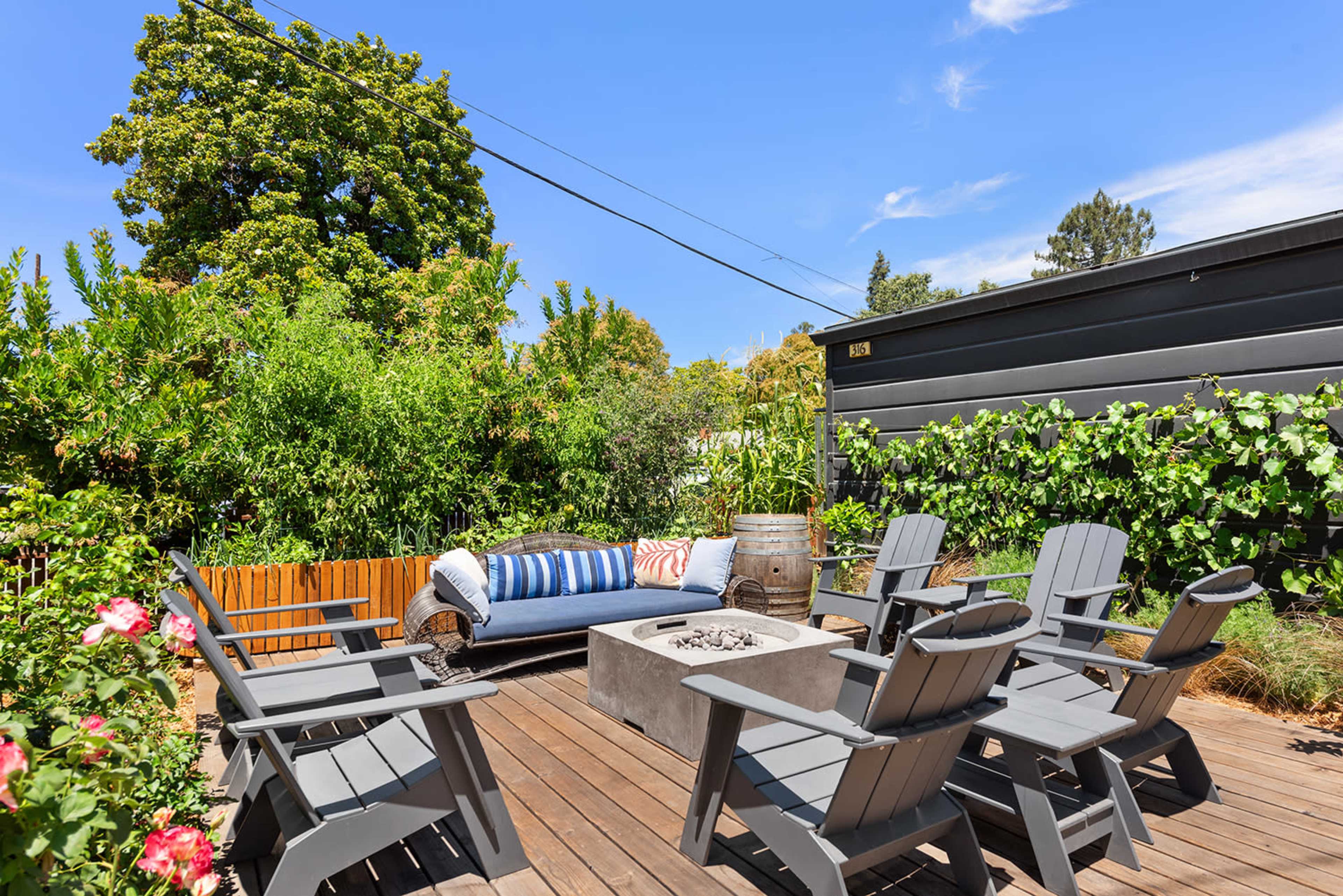 The image shows a wooden deck with a rectangular fire pit surrounded by several gray Adirondack chairs and a blue sofa, set amidst lush greenery.