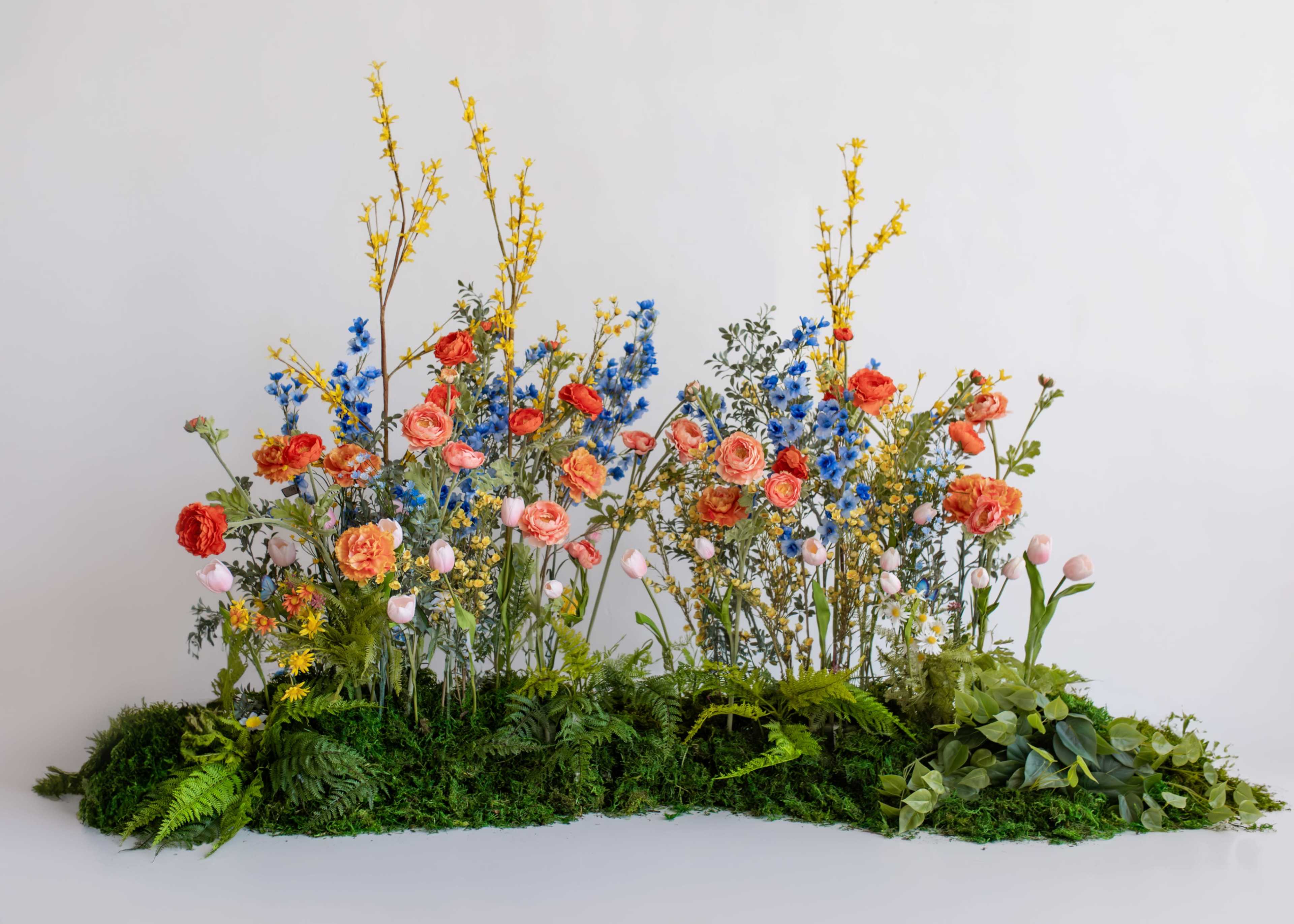 A vibrant arrangement of various flowers, including roses and delphiniums, is displayed against a white background, with greenery and moss at the base.