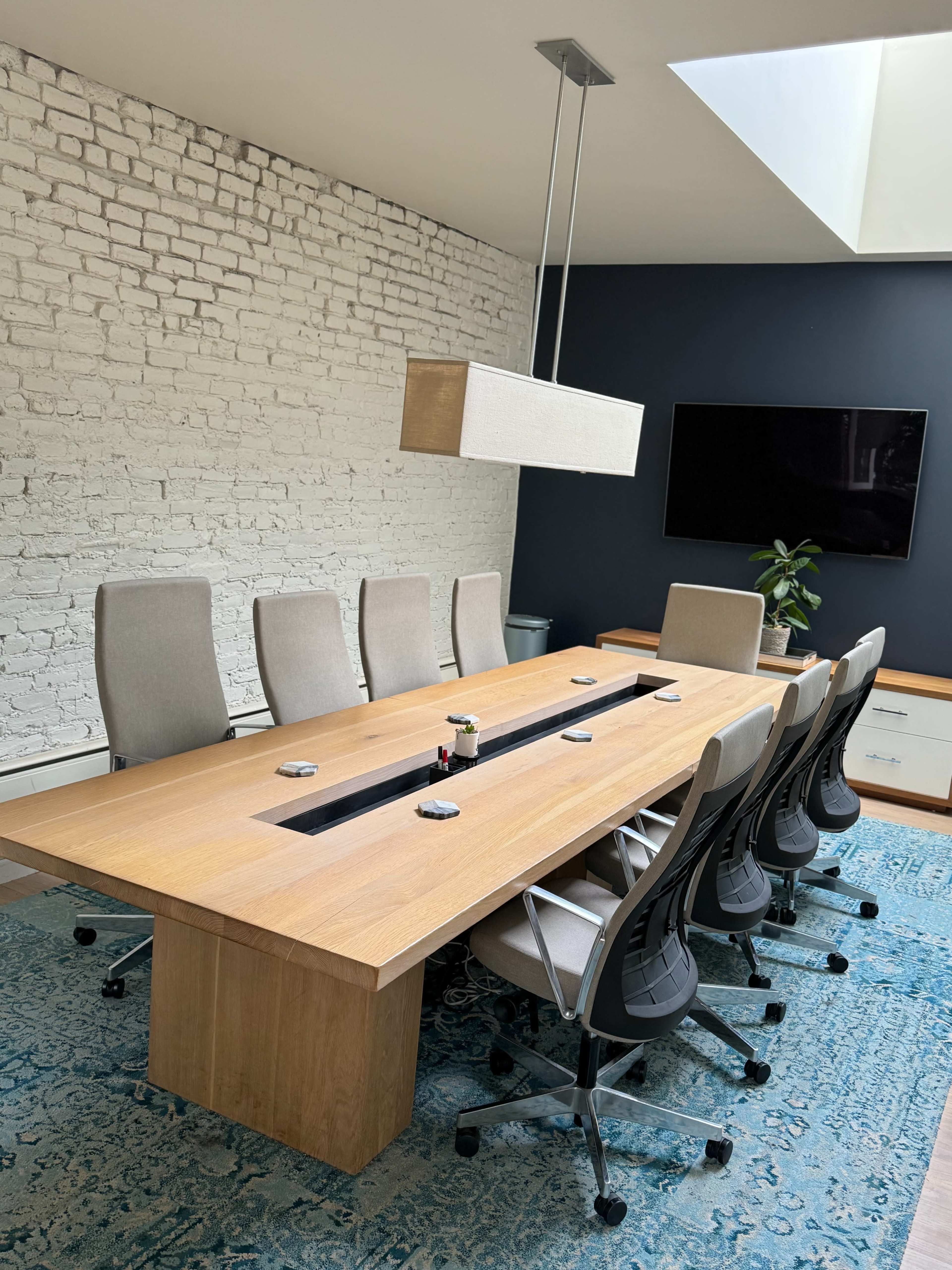 A modern conference room features a large wooden table surrounded by ergonomic chairs, with a skylight above and a television mounted on the wall.