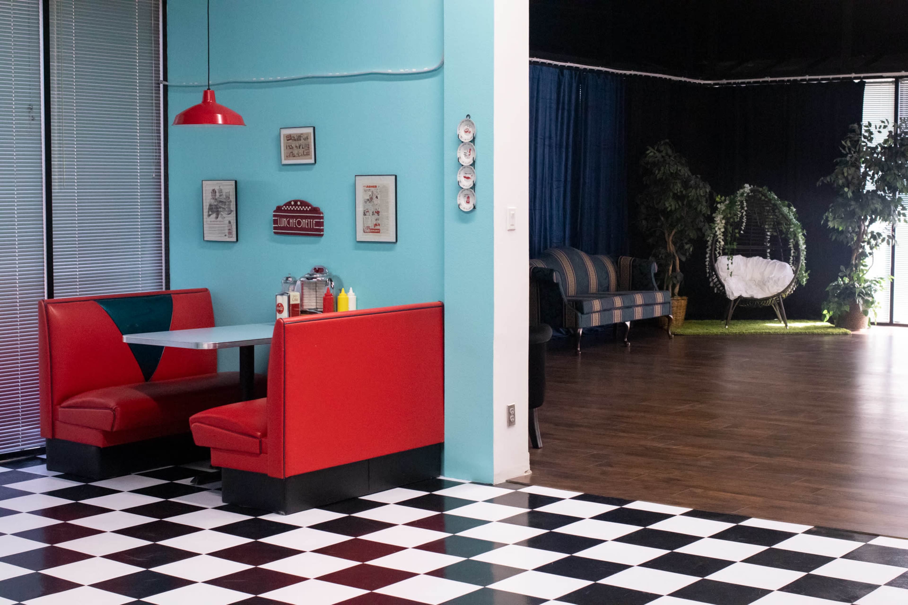 The image shows a retro-style diner booth in red with a checkered floor, adjacent to a seating area with a swing chair and plants.