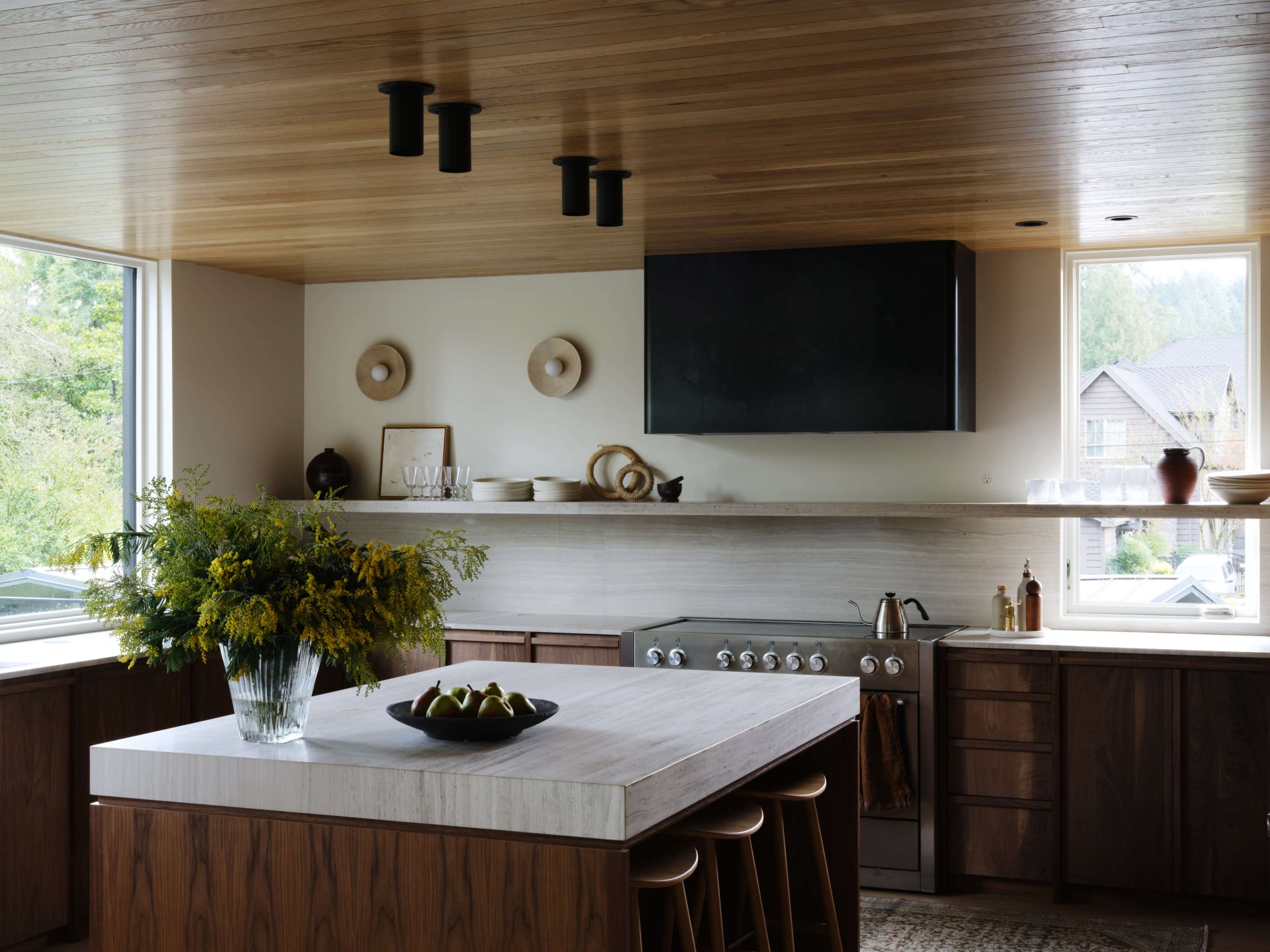 A modern kitchen features a wooden ceiling, sleek cabinetry, and a large island with a vase of flowers and a bowl of fruit.