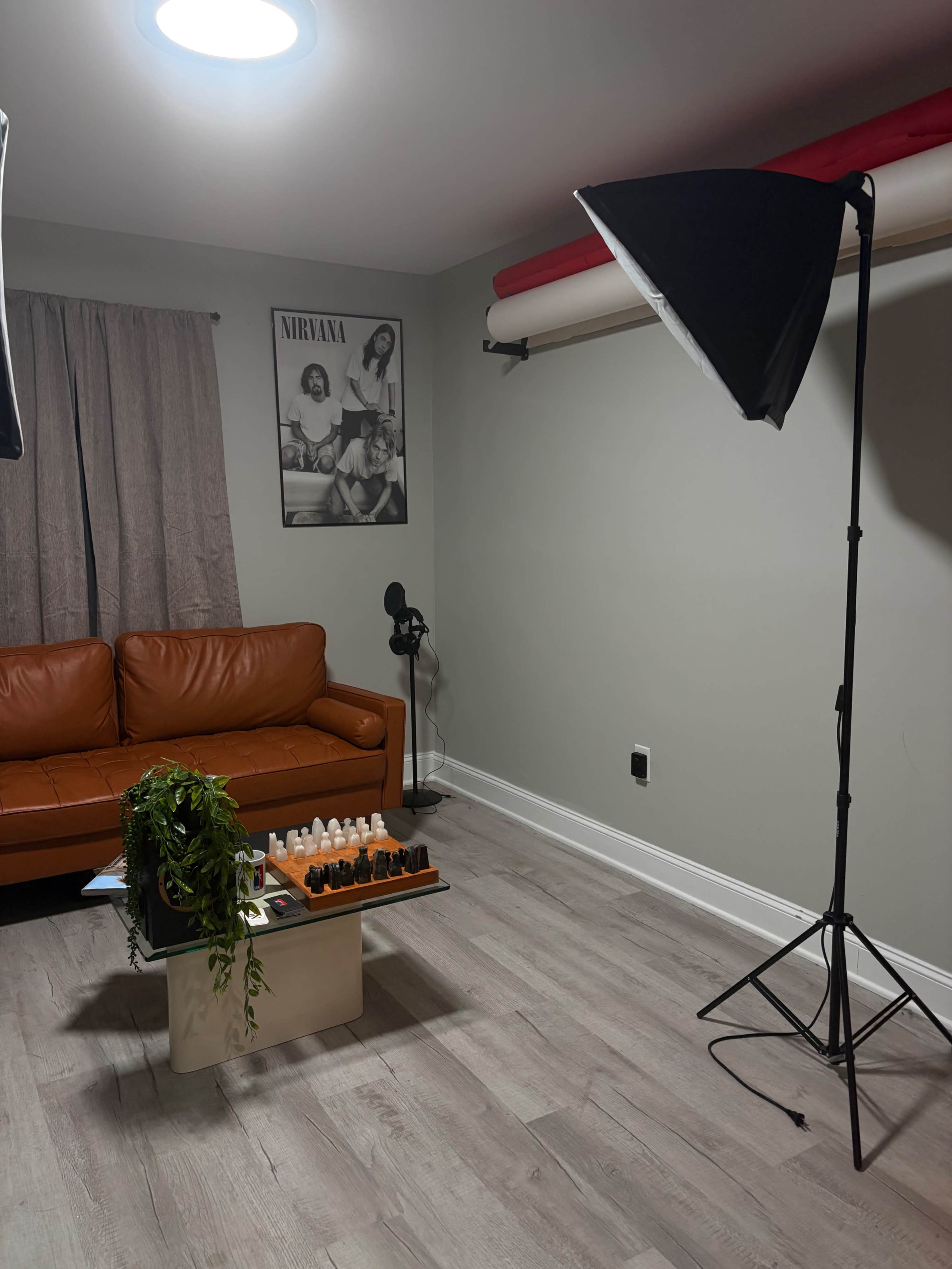 The image shows a room featuring a brown leather couch, a table with various bottles, and two softbox lights positioned nearby, with a poster of a band on the wall.