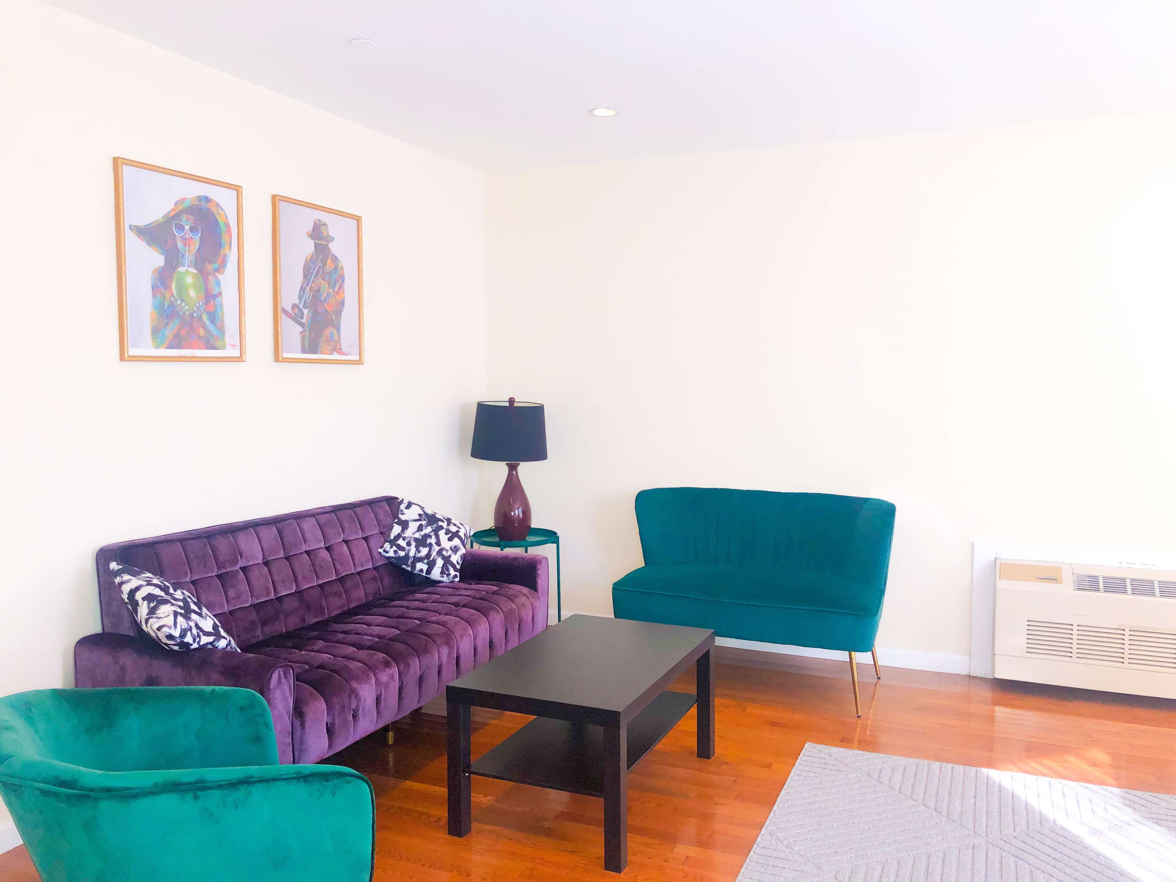 A well-lit living room with a purple couch, a green sofa, and a black coffee table, adorned with colorful artwork on the walls.