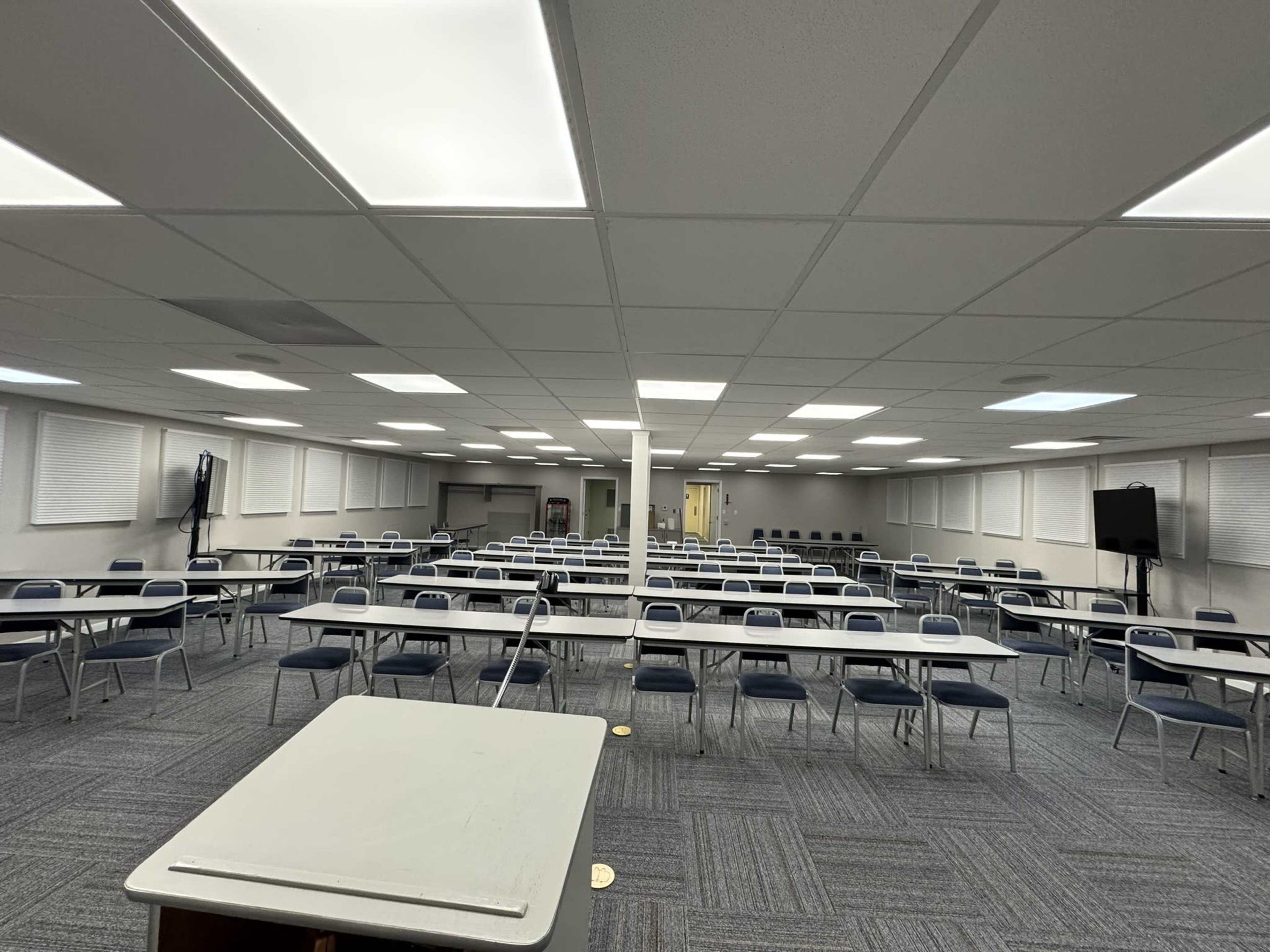 The image shows a large, empty classroom with rows of white tables and blue chairs arranged neatly on a gray carpet.