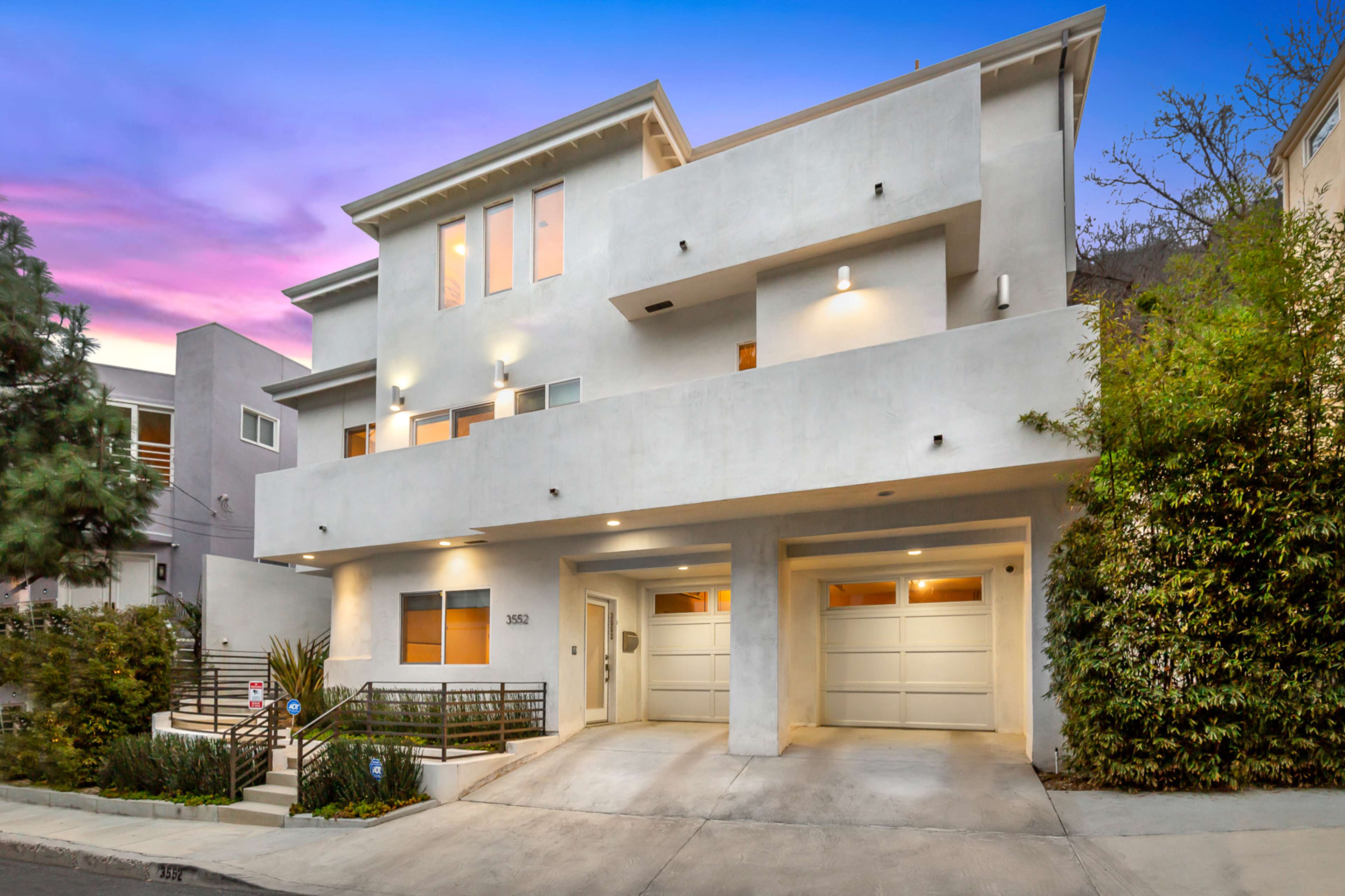 A modern three-story home with a smooth exterior, featuring two garage doors and landscaping, sits at the end of a driveway under a colorful sunset sky.