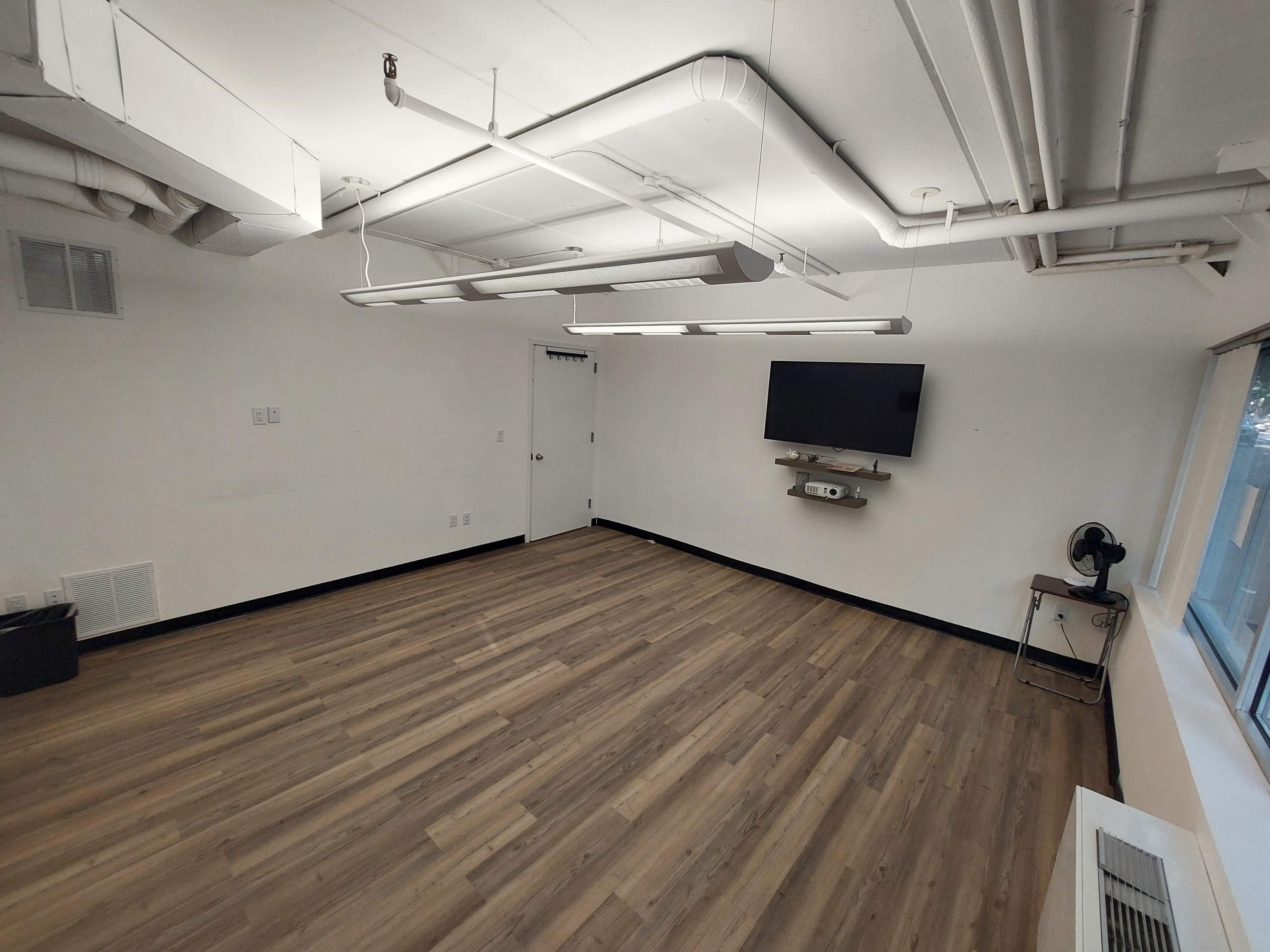 The image shows an empty room with wooden flooring, white walls, a large window, a television mounted on the wall, and a table with a fan in the corner.