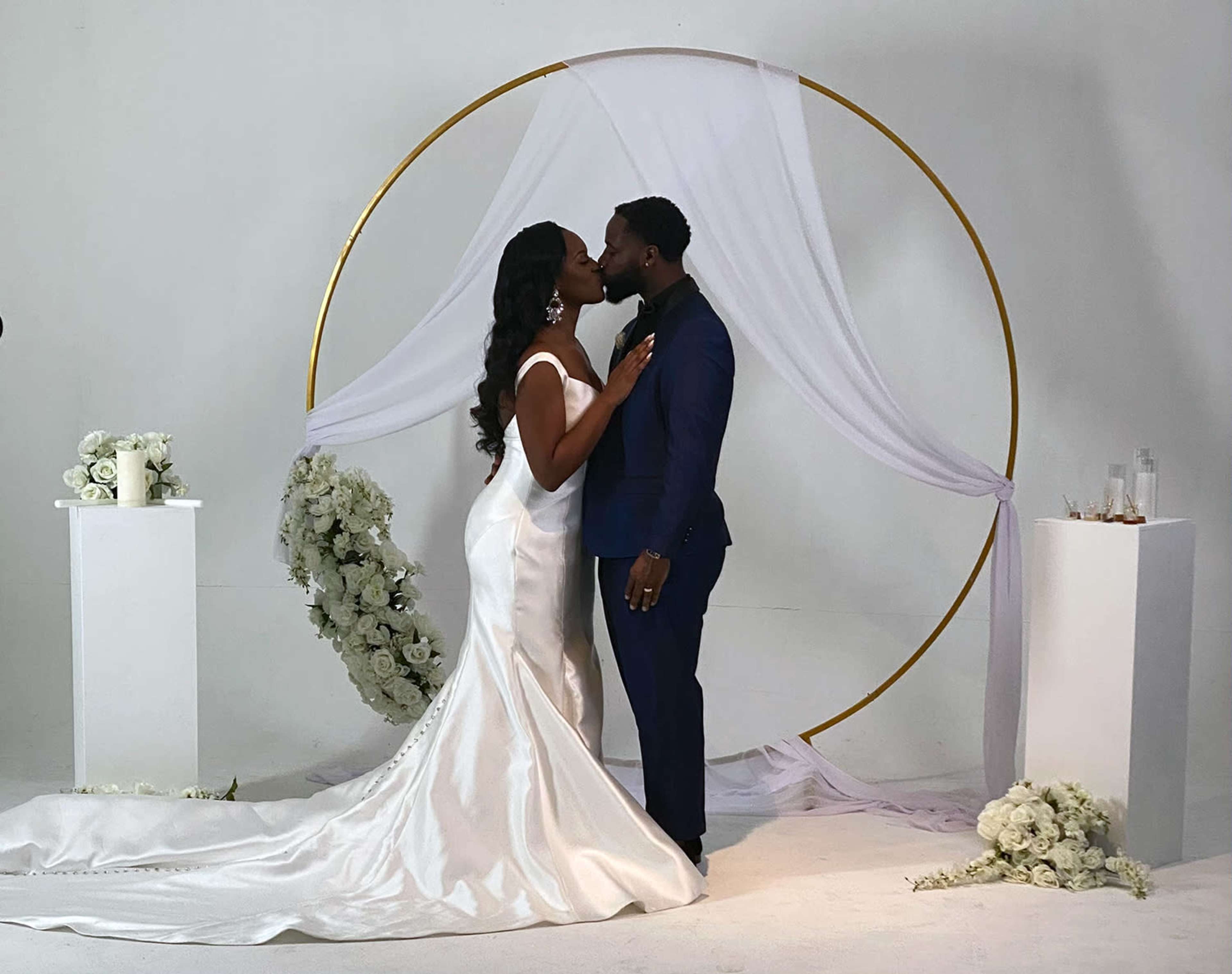 A bride and groom kiss in front of a floral archway draped with white fabric.