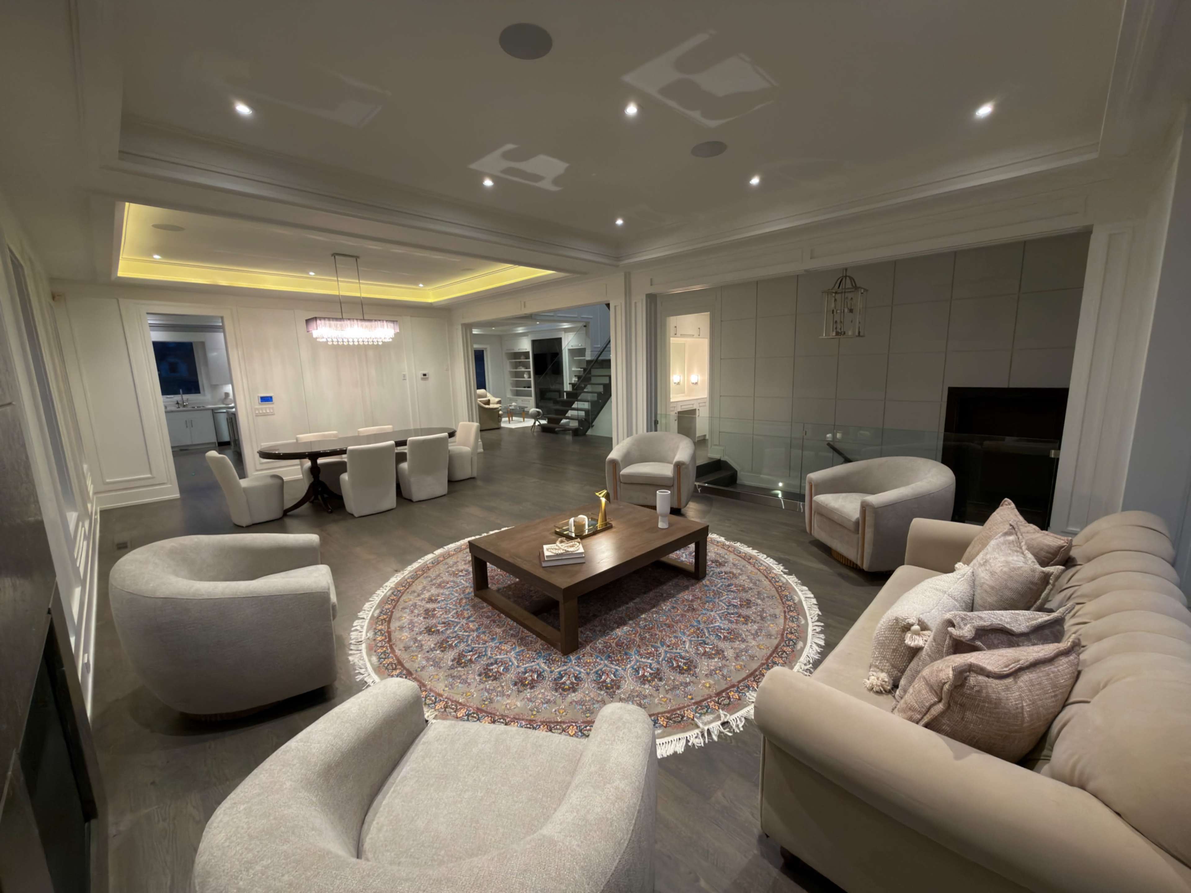 The image shows a modern living room with a circular area rug, comfortable seating, a coffee table, and a staircase leading to another level.