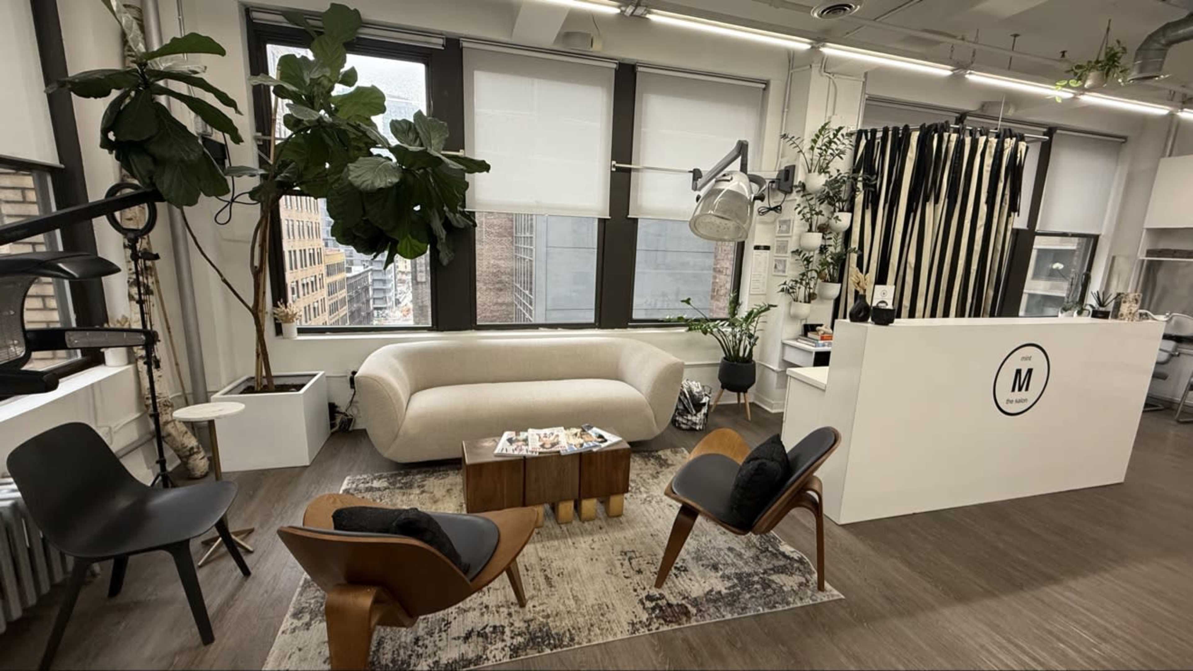 The image shows a modern office space featuring a light-colored couch, several plants, a round table with snacks, and a reception area with a logo.