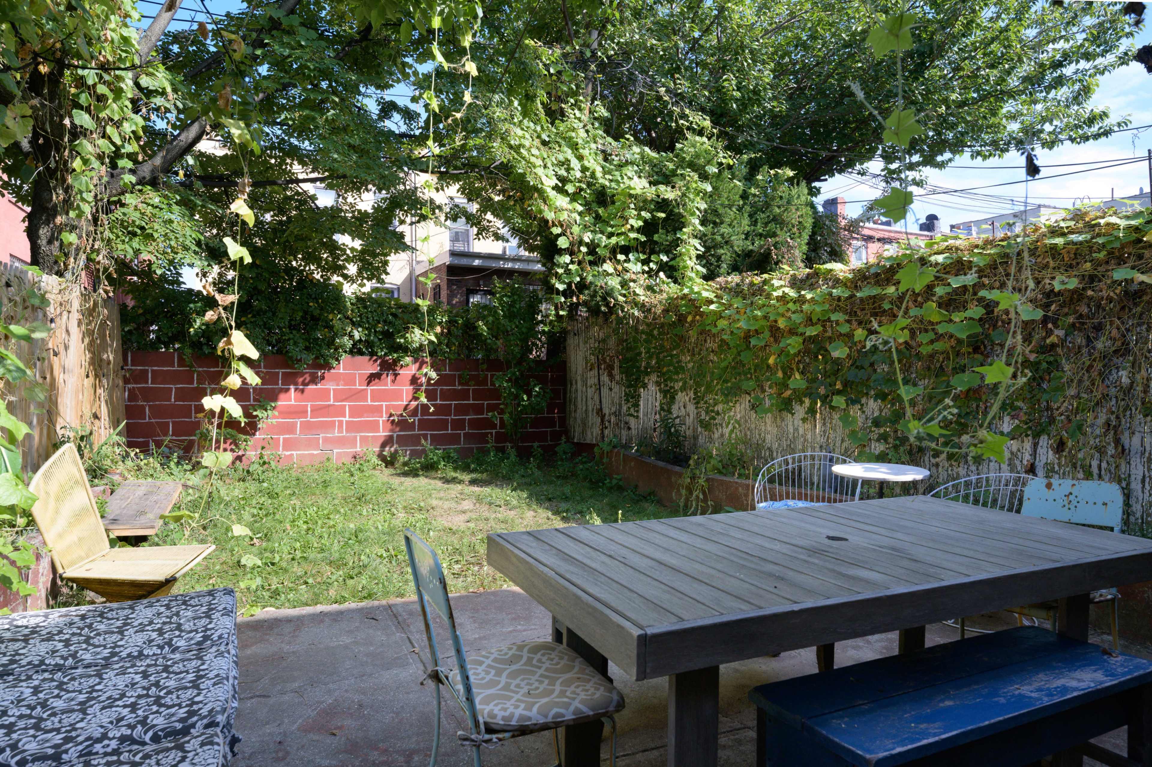 The image shows a small, overgrown backyard surrounded by wooden fences, with a wooden table and several chairs arranged in the center.
