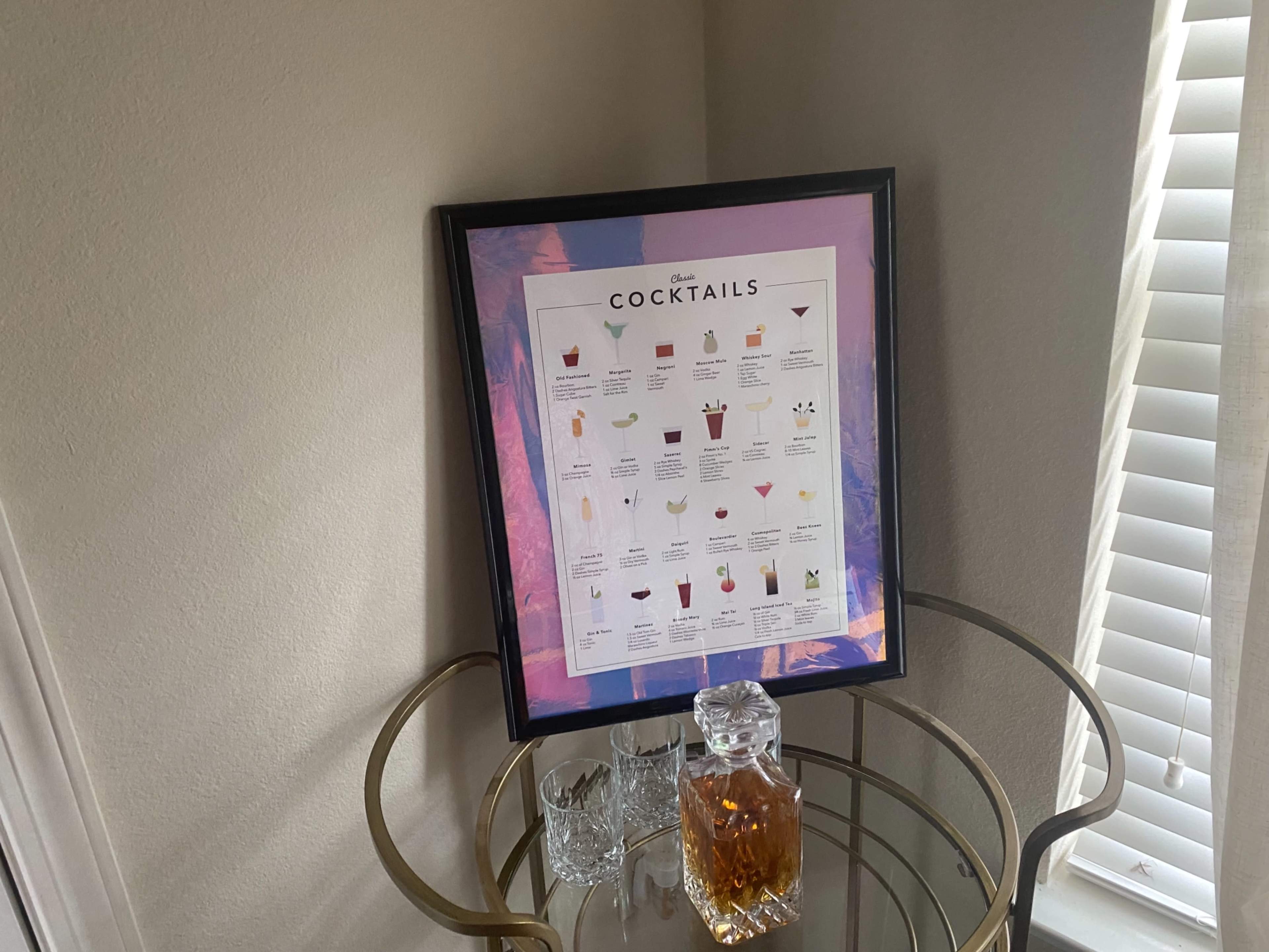 A framed cocktail recipe poster is displayed on a wall above a decorative bar cart with a glass decanter and two empty glasses.