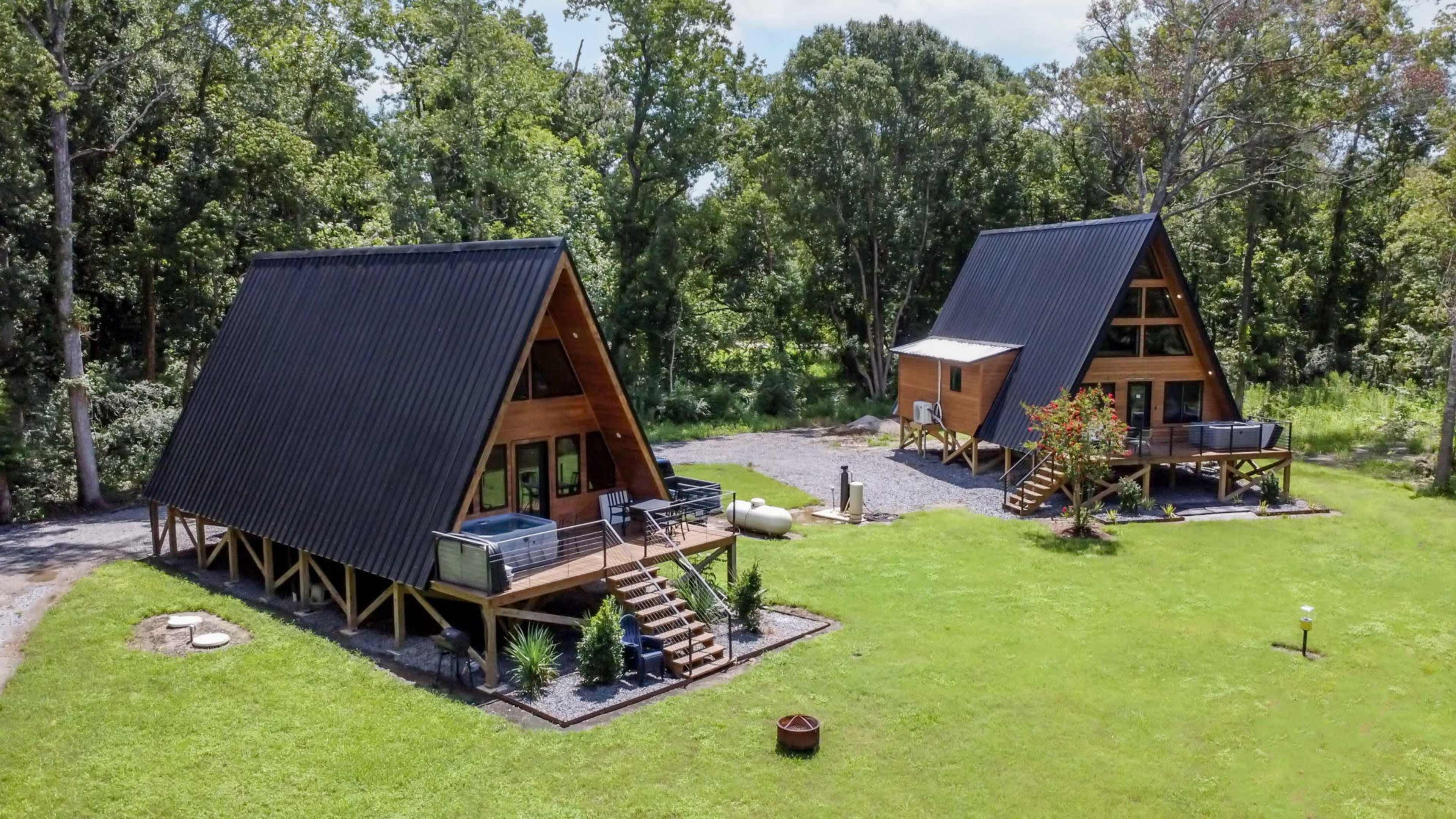Two A-frame cabins with black roofs are situated on a grassy area surrounded by trees.