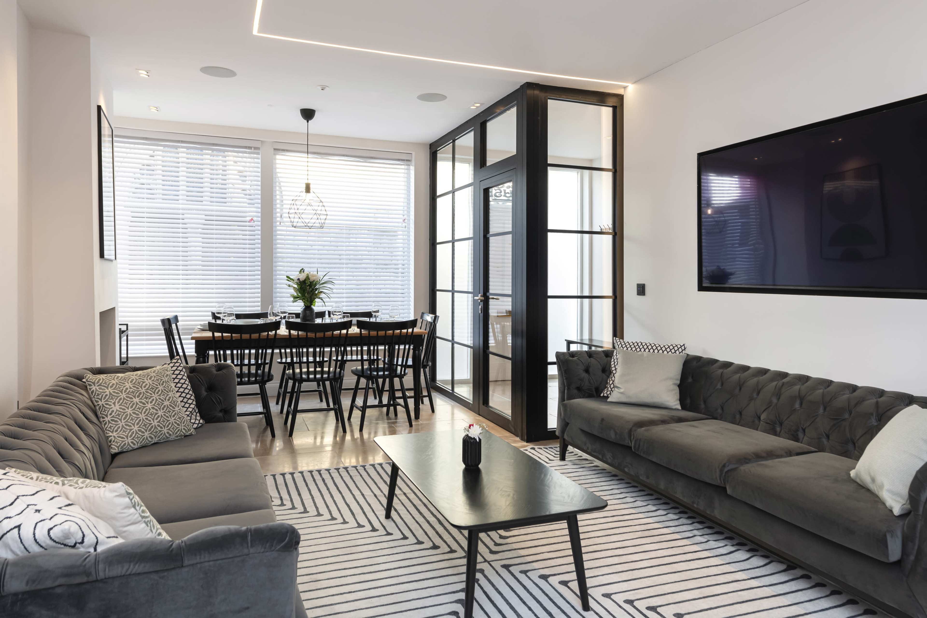 A modern living room featuring two gray sofas, a black coffee table on a striped rug, and a large flat-screen TV mounted on the wall, with a dining area visible through glass doors.