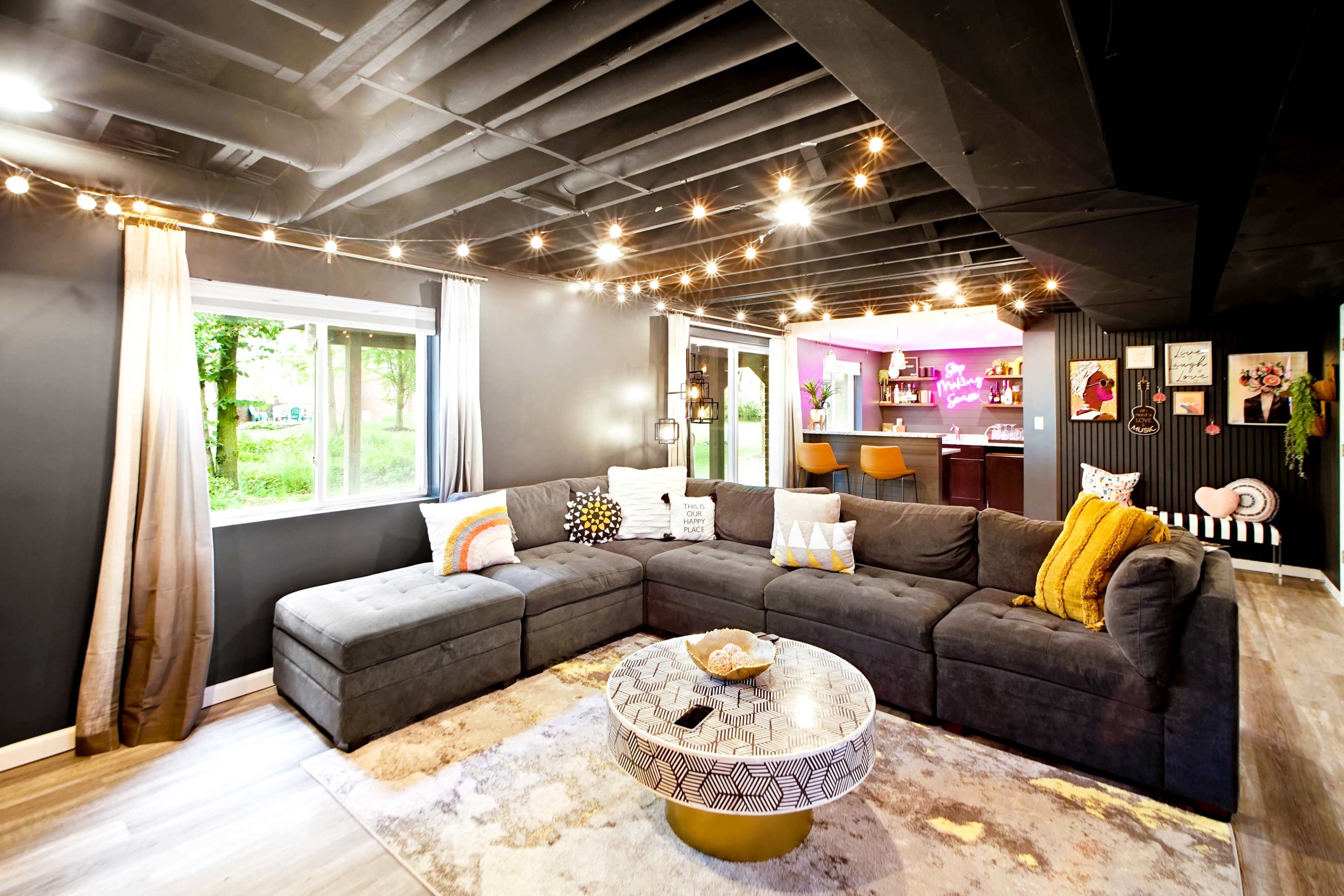 The image shows a modern living room with a large sectional sofa, a round coffee table, and a bar area illuminated by colorful lights.
