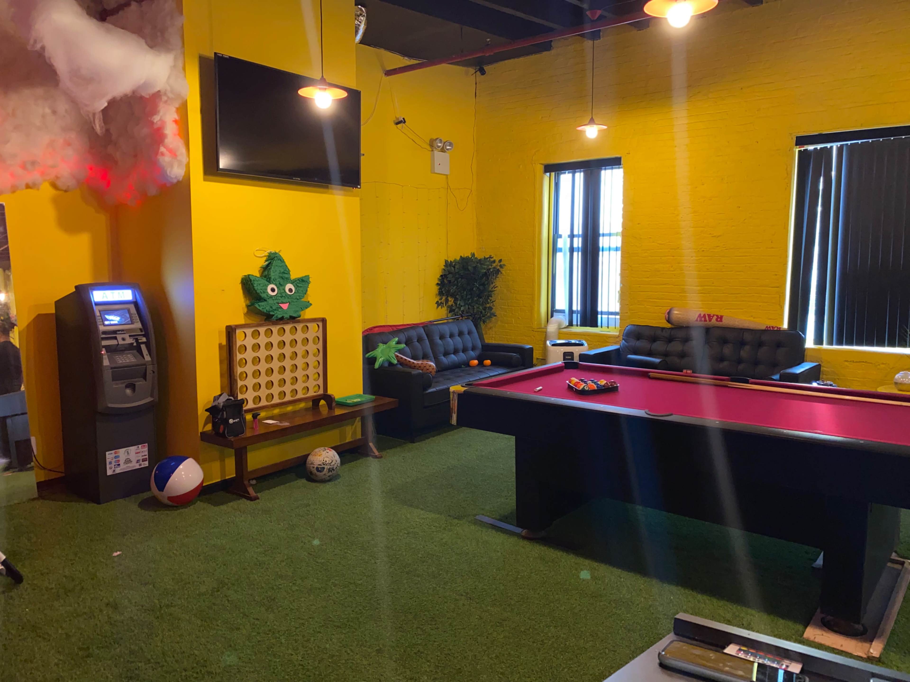 The scene features a recreational room with a pool table, two black couches, an ATM, a large yellow wall, and artificial grass flooring.