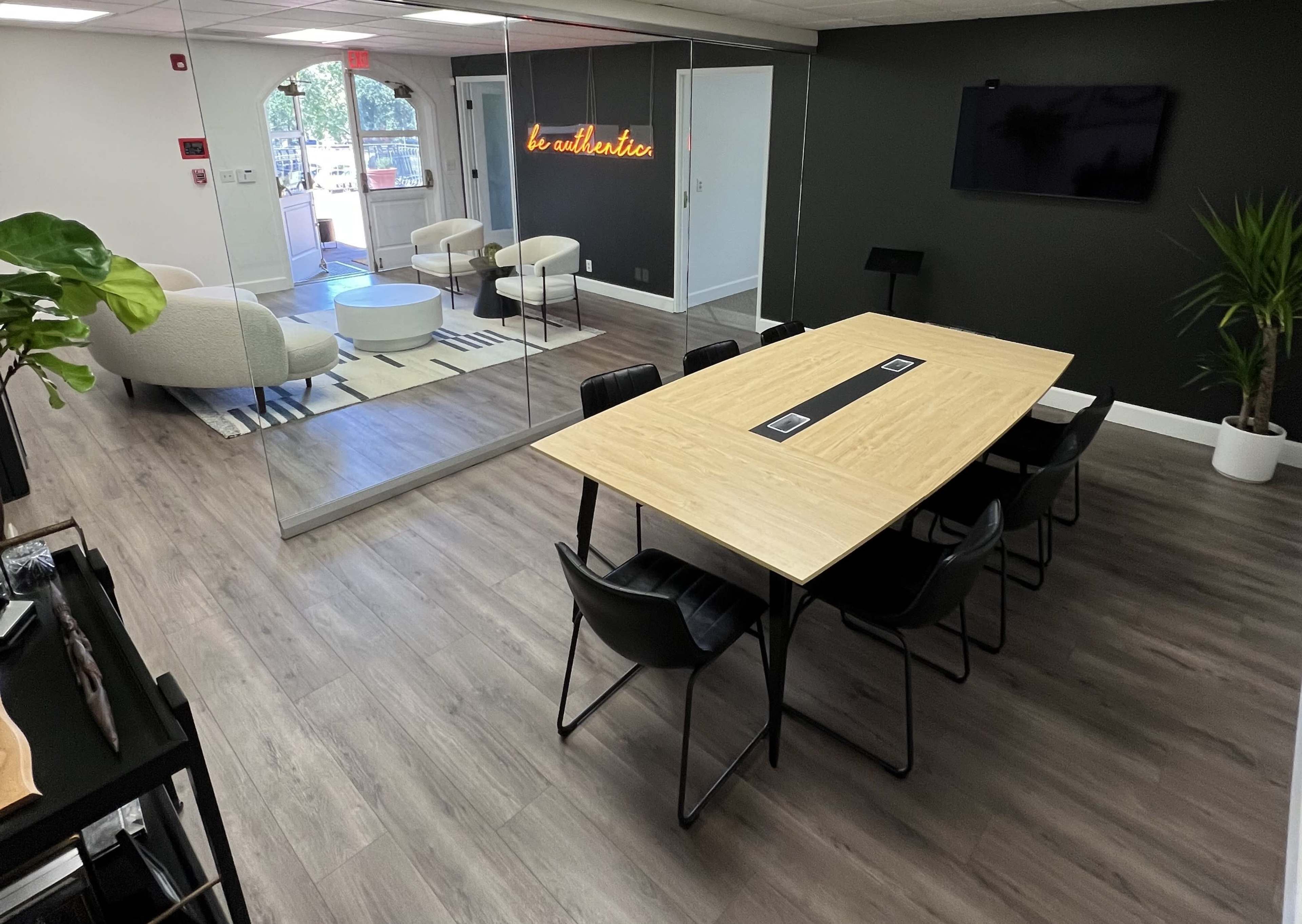 A modern office space with a large wooden table surrounded by black chairs, a seating area with white chairs, and a wall featuring a neon sign that reads "be authentic."