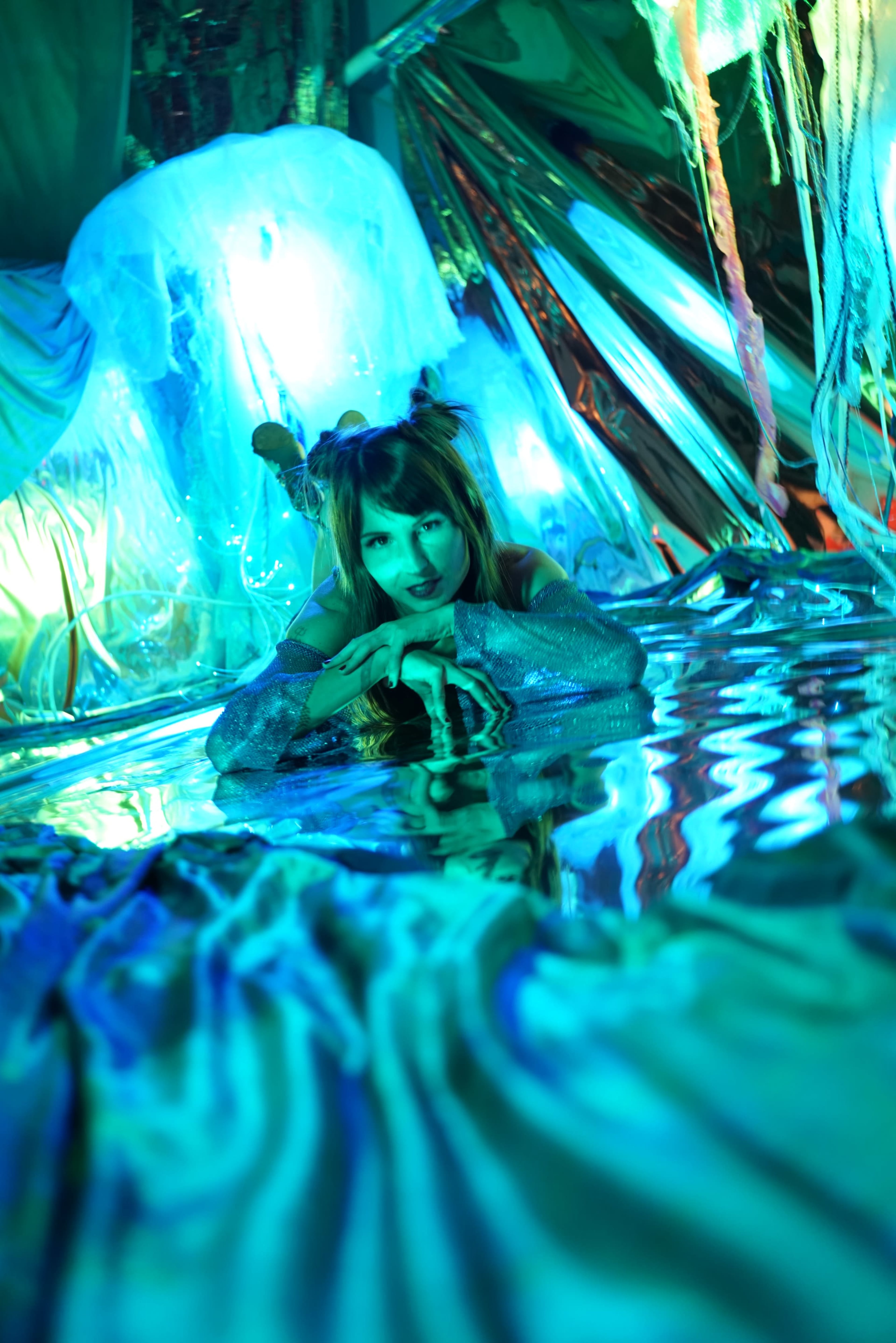 A person with hair styled in playful buns lies on a reflective surface surrounded by colorful, illuminated drapery.