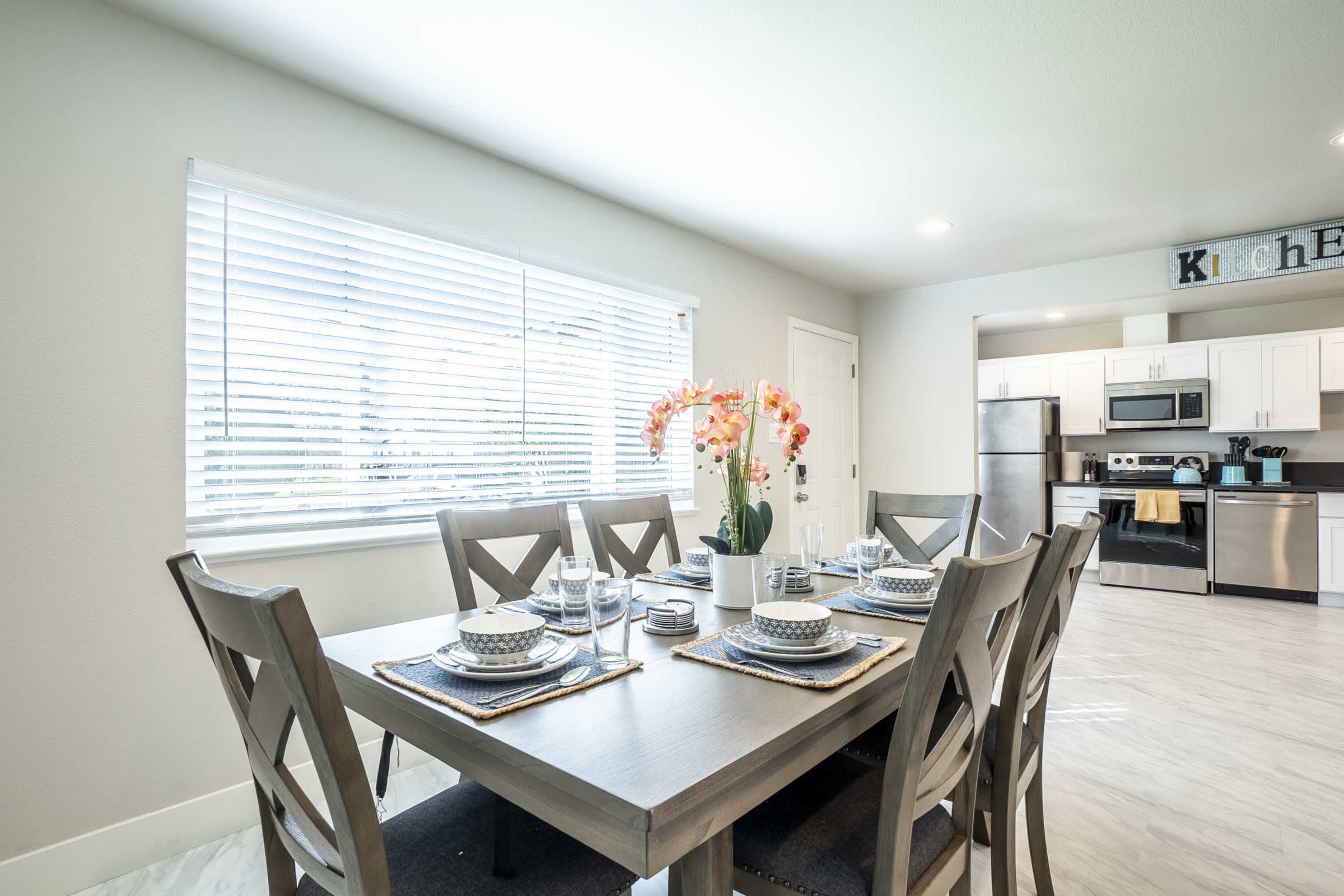 A dining room is set with a wooden table, complete with plates, glasses, and a vase of flowers, with a window providing natural light.