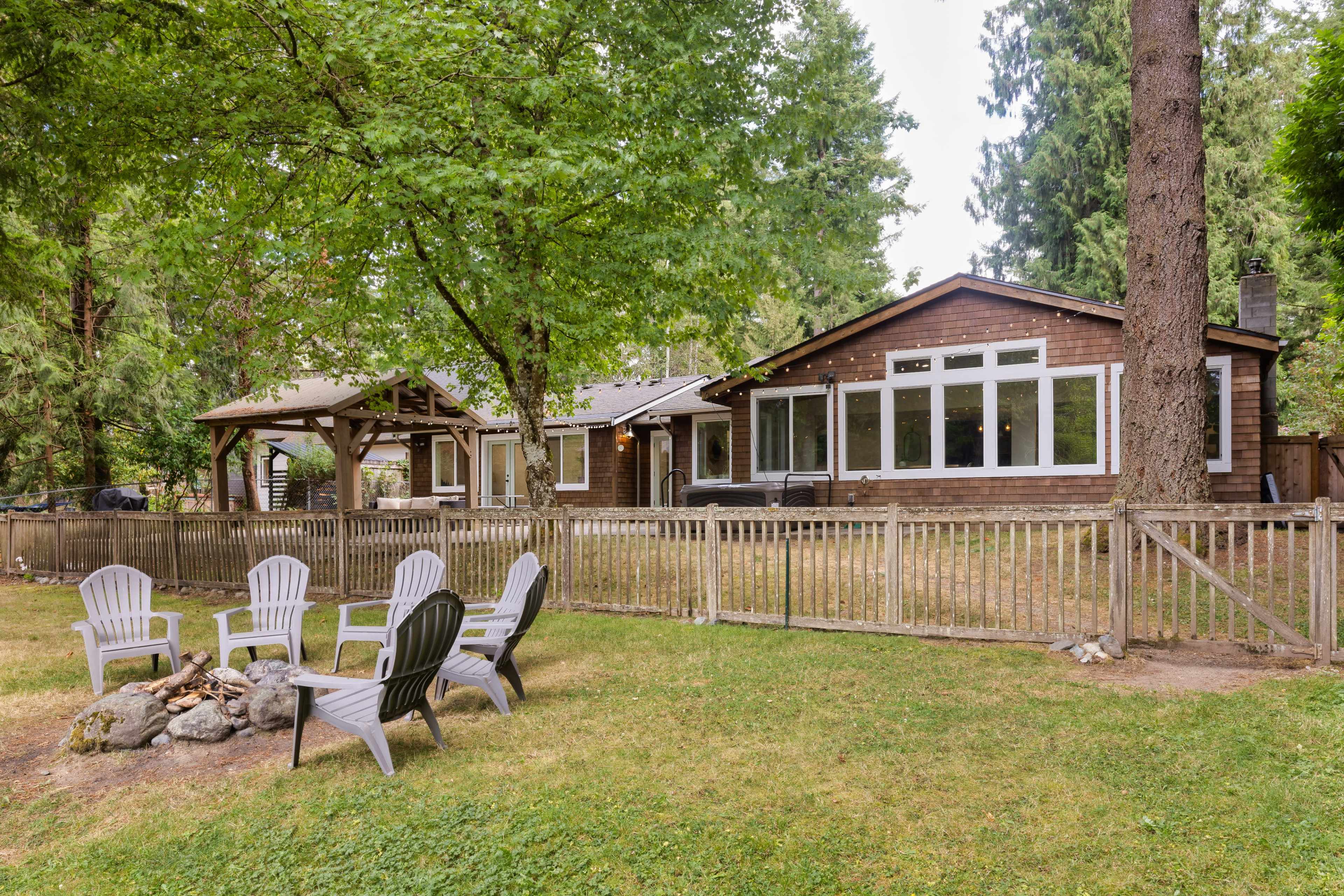 A wooden house with large windows is surrounded by trees and features a fenced yard with a fire pit and several Adirondack chairs.