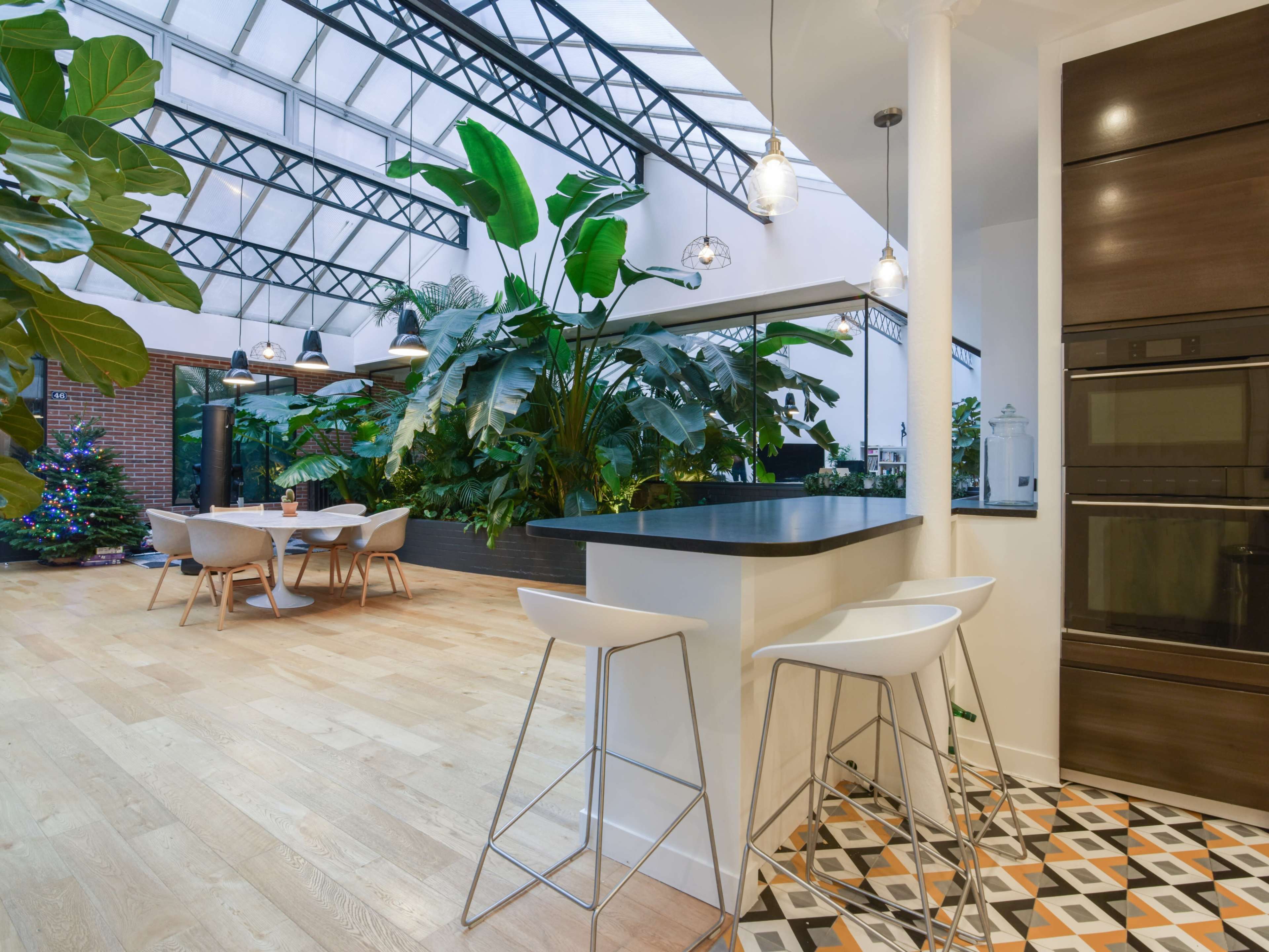 The image shows a modern kitchen area with a bar counter and stools, leading into a spacious dining area filled with greenery under a glass ceiling.