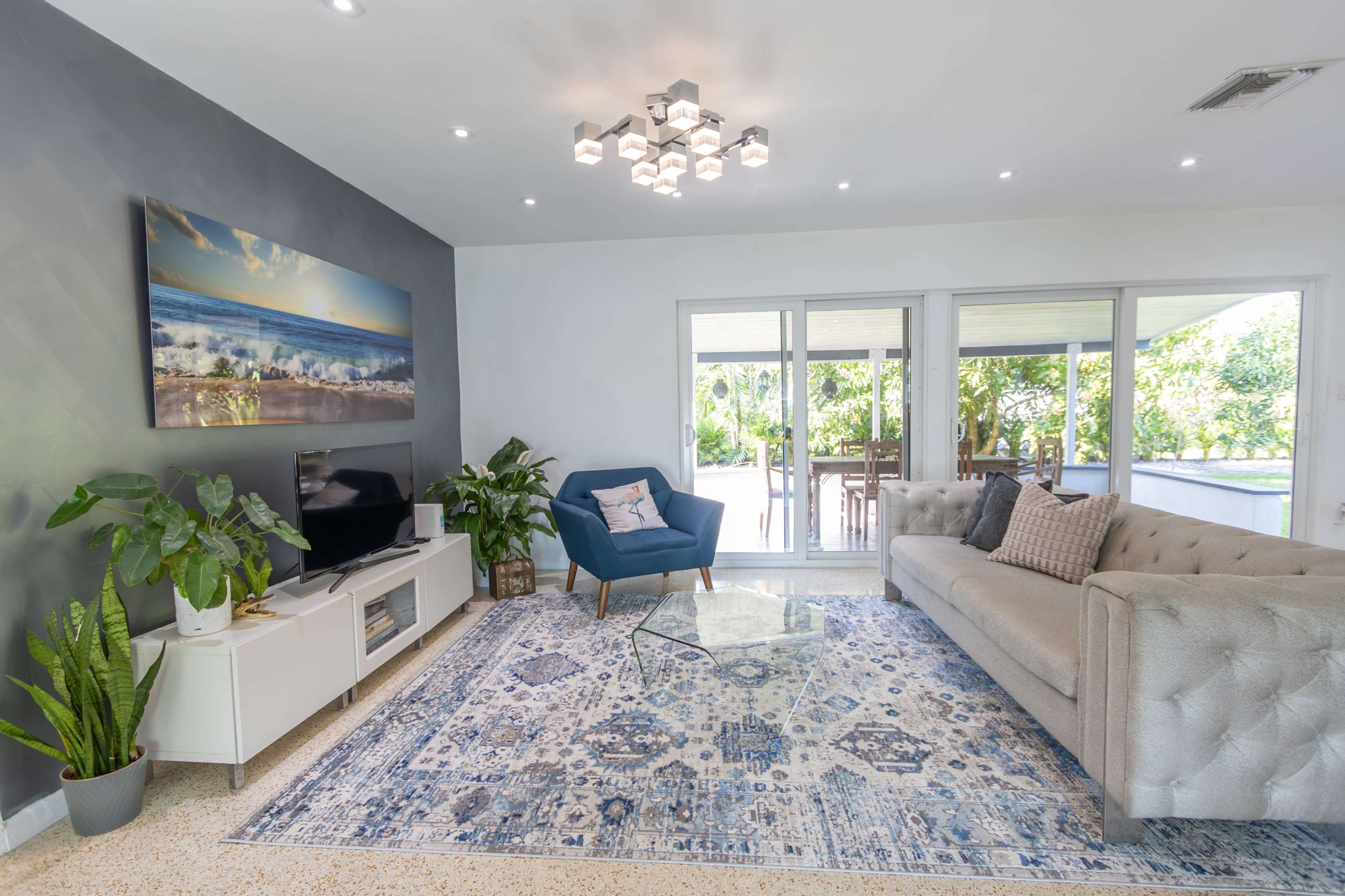 A modern living room featuring a light gray sofa, a blue accent chair, a glass coffee table, a large area rug, and a television against a wall decorated with a landscape painting.
