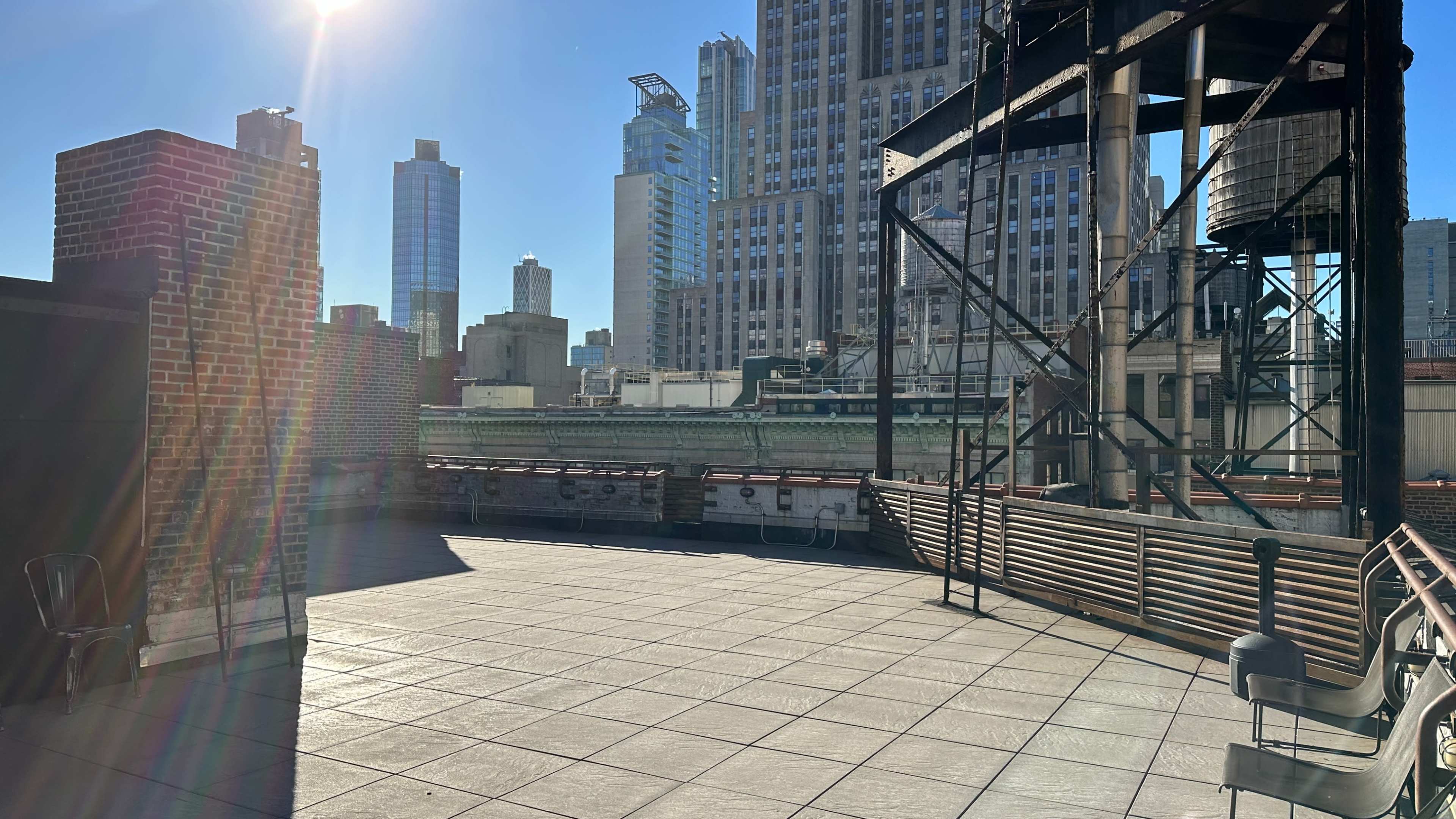 The image shows a rooftop terrace with modern city buildings in the background under a clear blue sky.