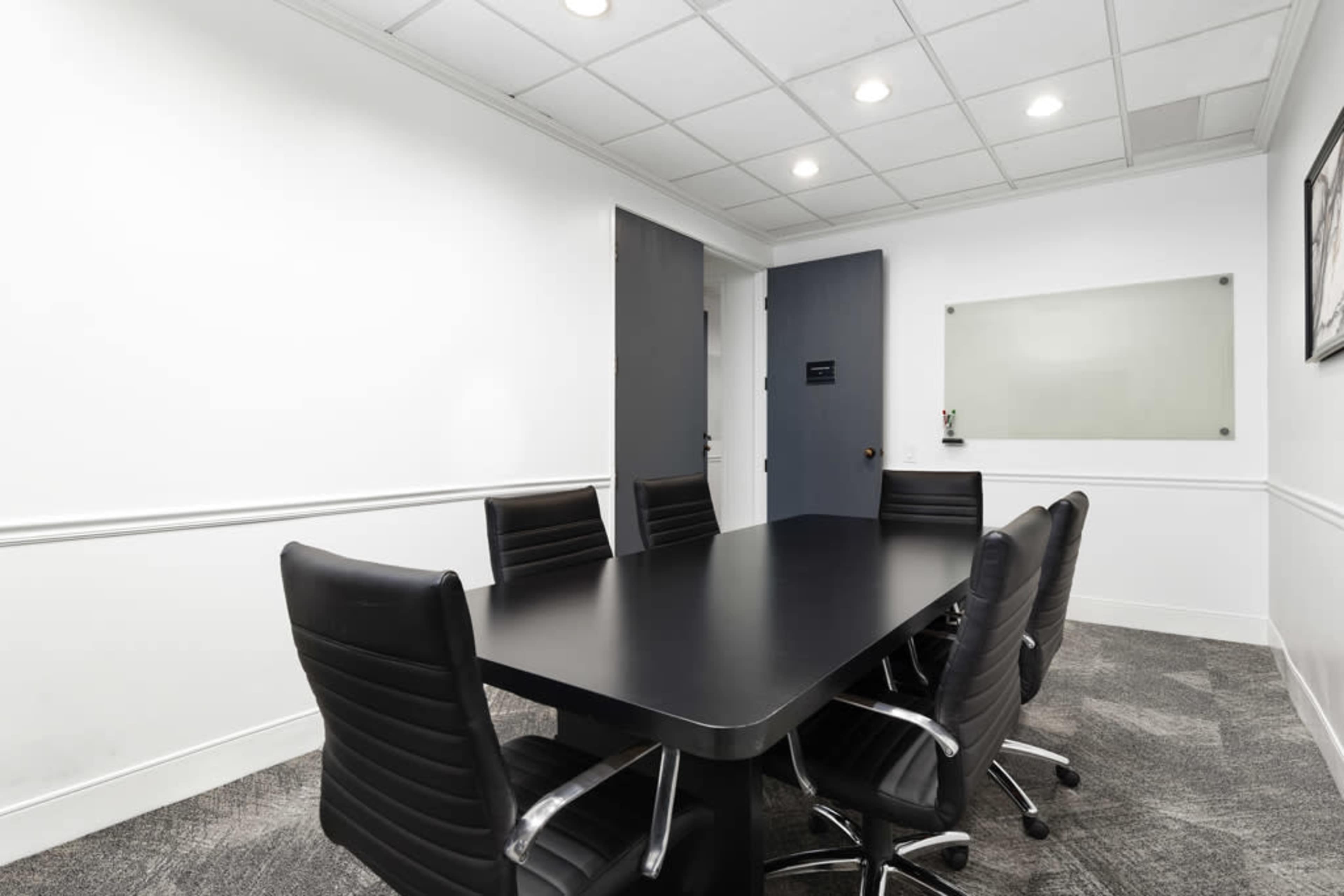 The image shows a modern conference room with a black table and six black chairs, against a white wall with a glass board and a door.