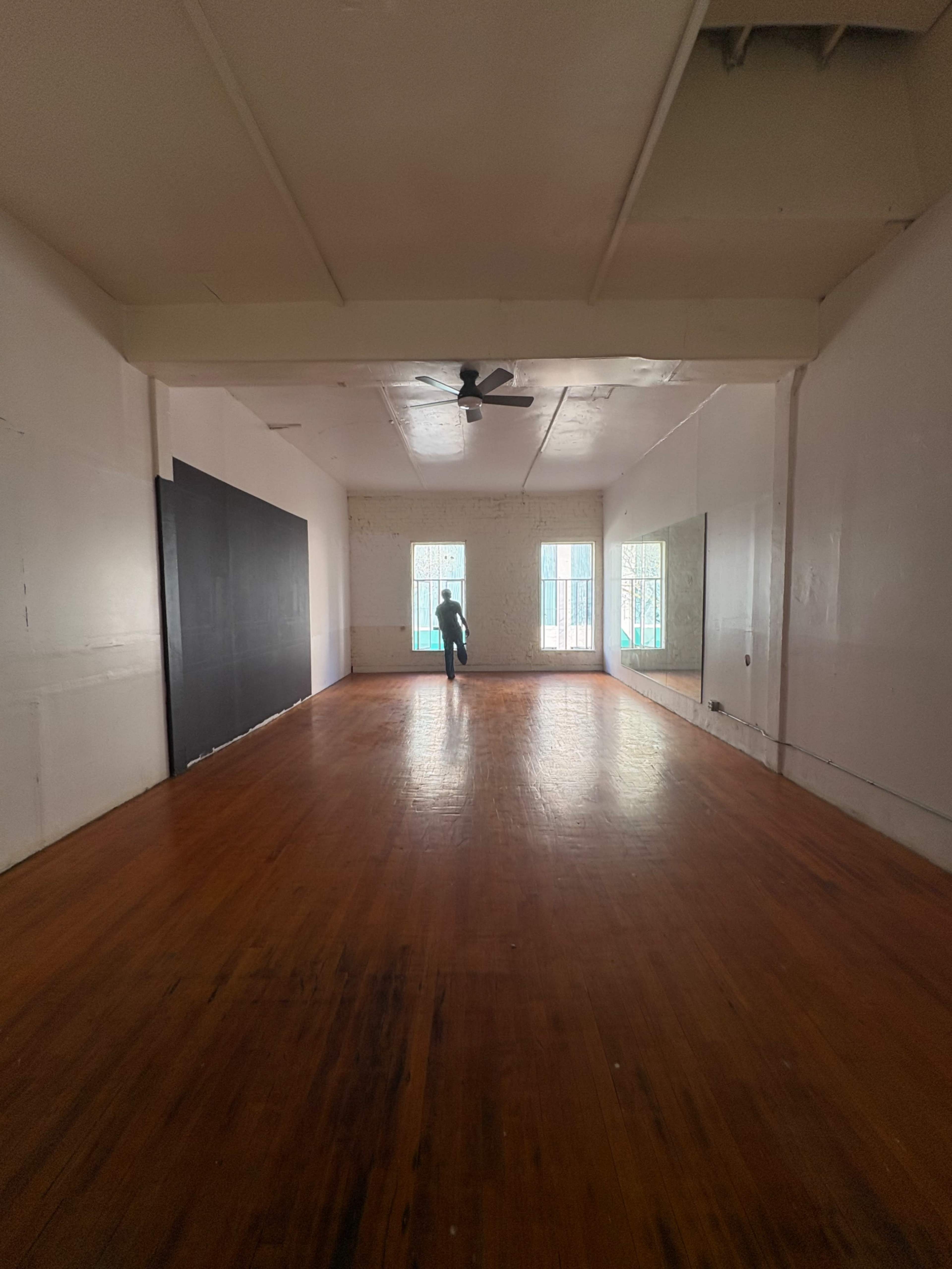 A person is standing in a spacious room with wooden floors and three large windows, casting natural light onto the empty space.