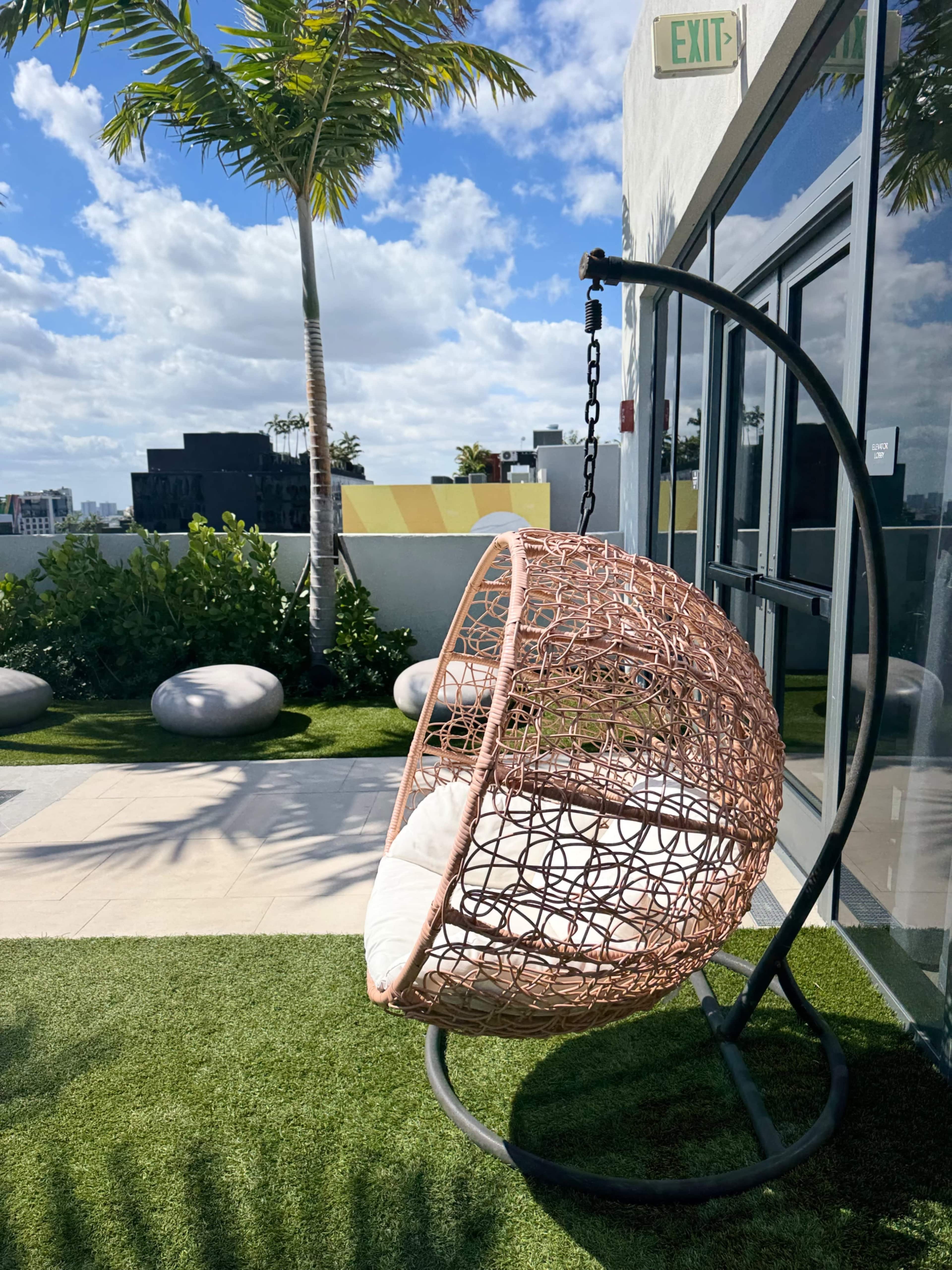 A large, round, wicker swing chair hangs from a metal frame on a green lawn near a building with large windows.