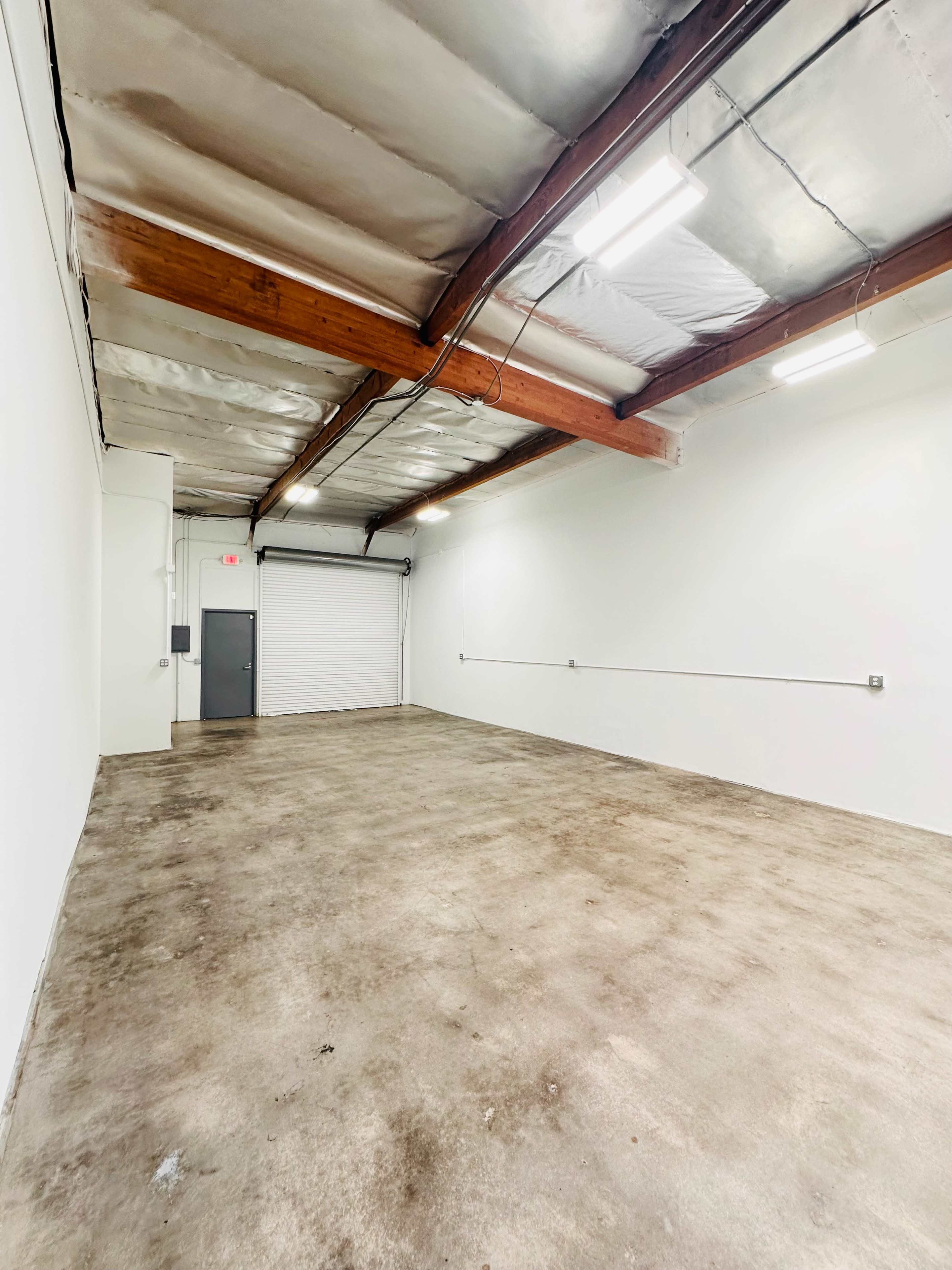 An empty industrial space with concrete flooring, exposed wooden beams, and a roll-up garage door.