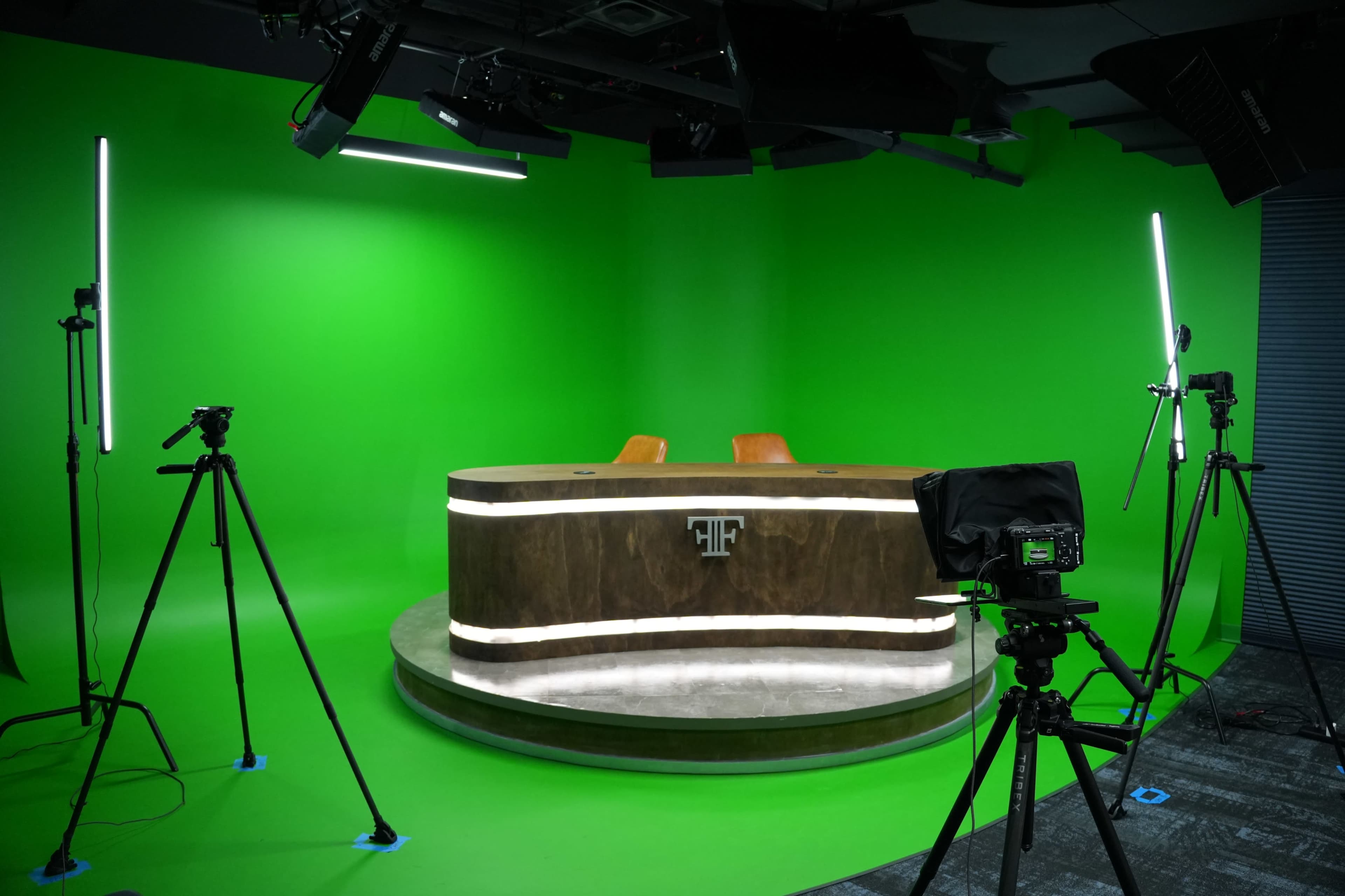 The image shows a television studio setup with a wooden news desk, two orange chairs, and a green backdrop, surrounded by tripods and lighting equipment.
