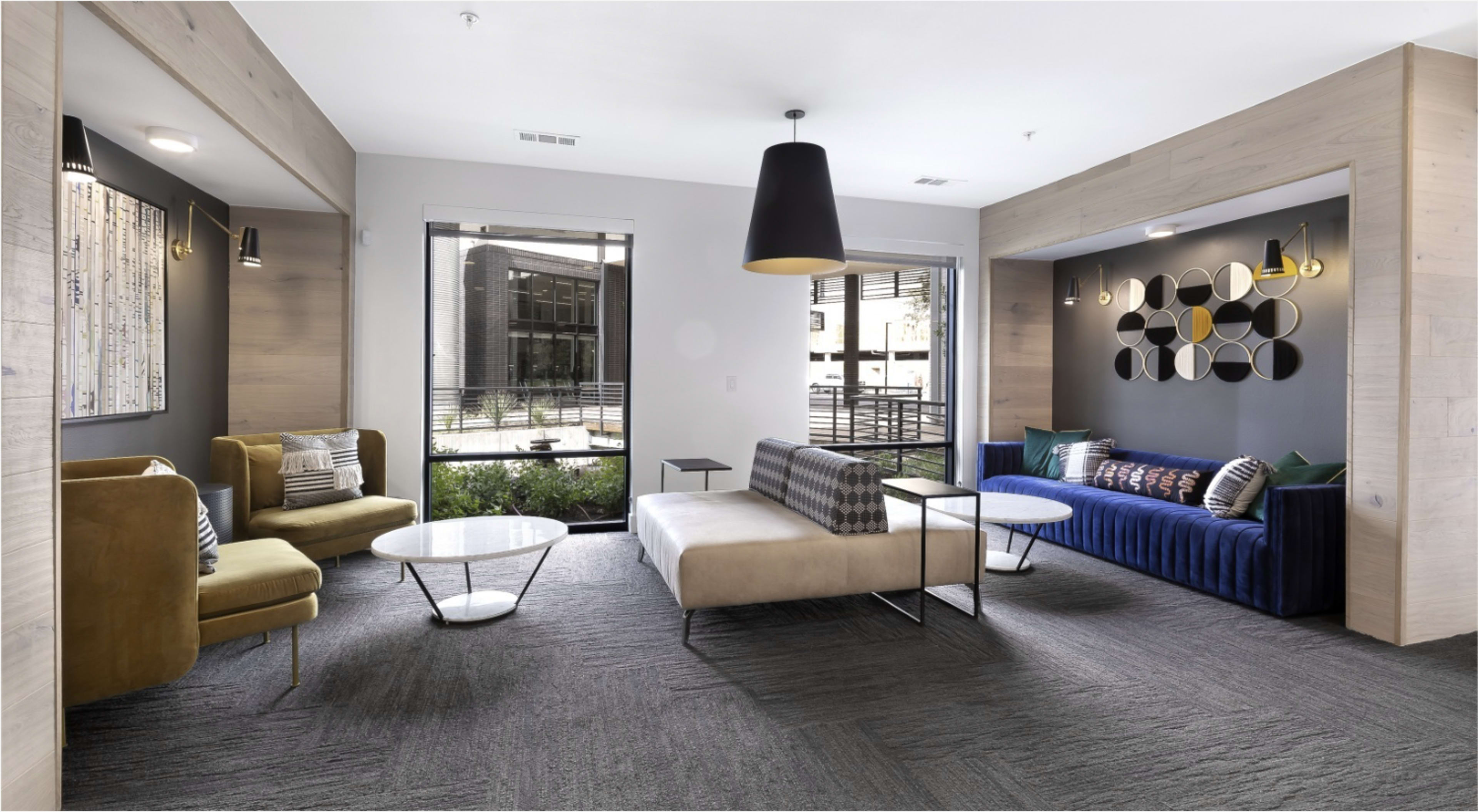 The image shows a modern lounge area featuring a mix of seating options, including a beige sofa and colorful armchairs, with large windows providing natural light.