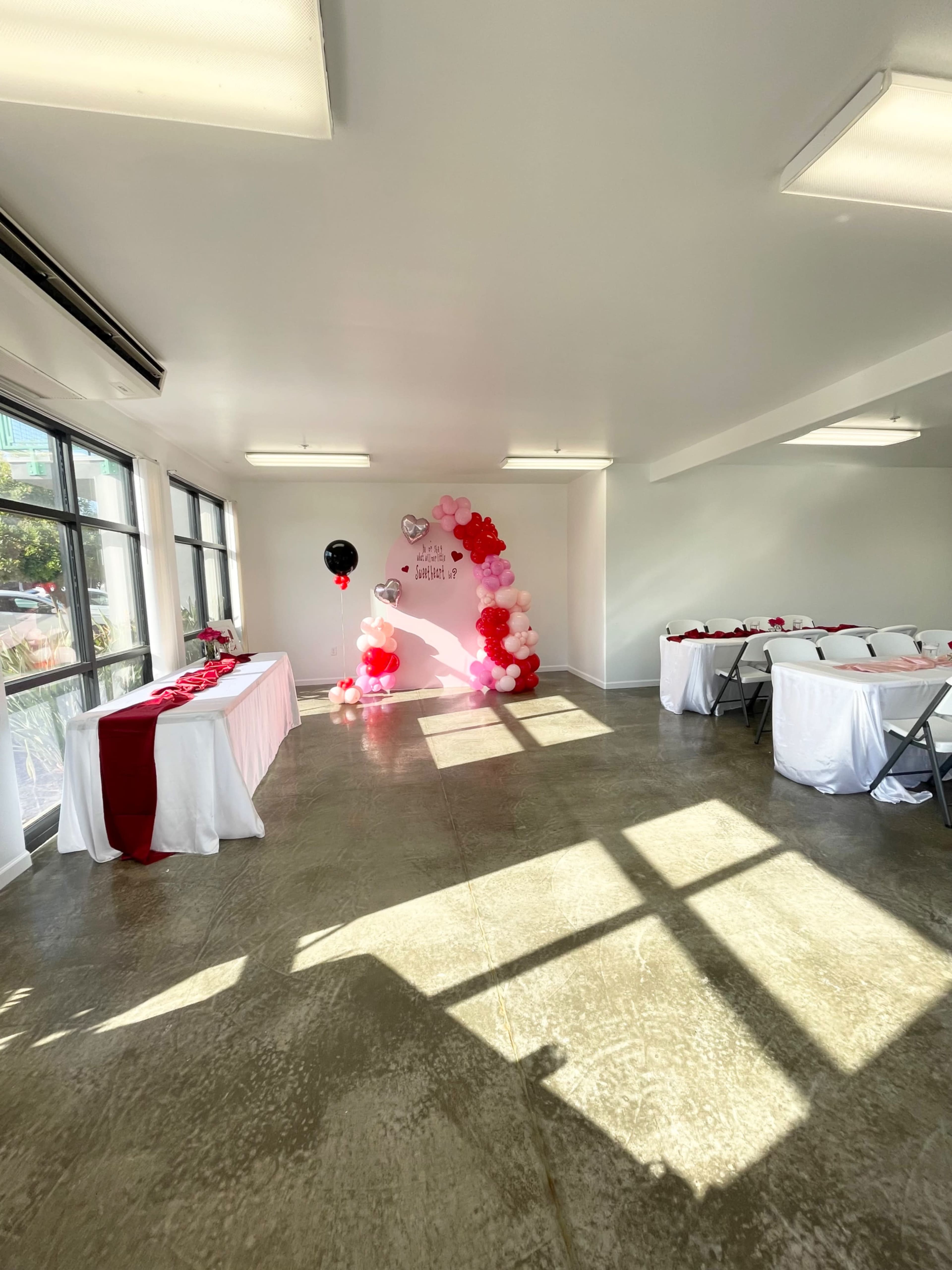 A brightly lit event space features tables covered in white cloth with red accents, and a decorative balloon arch in pink and white against the wall.