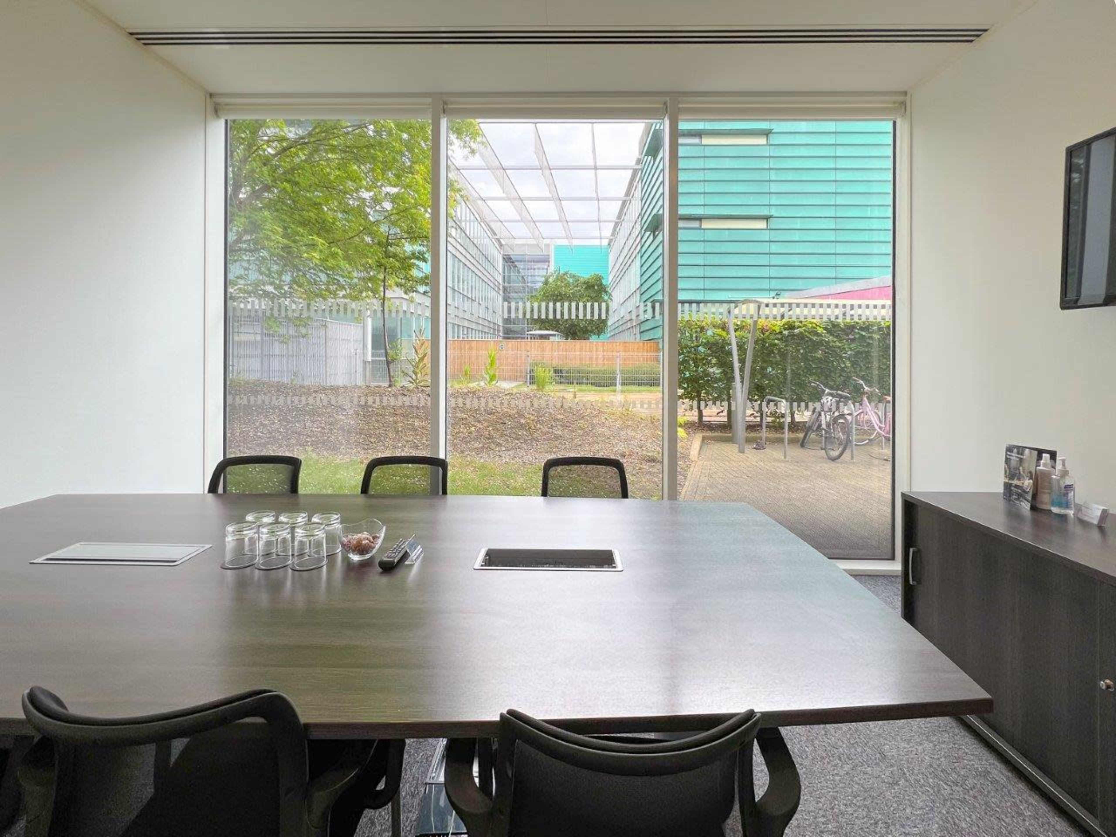 The image shows a modern conference room with a large table and glass walls overlooking an outdoor area with green trees and buildings in the background.