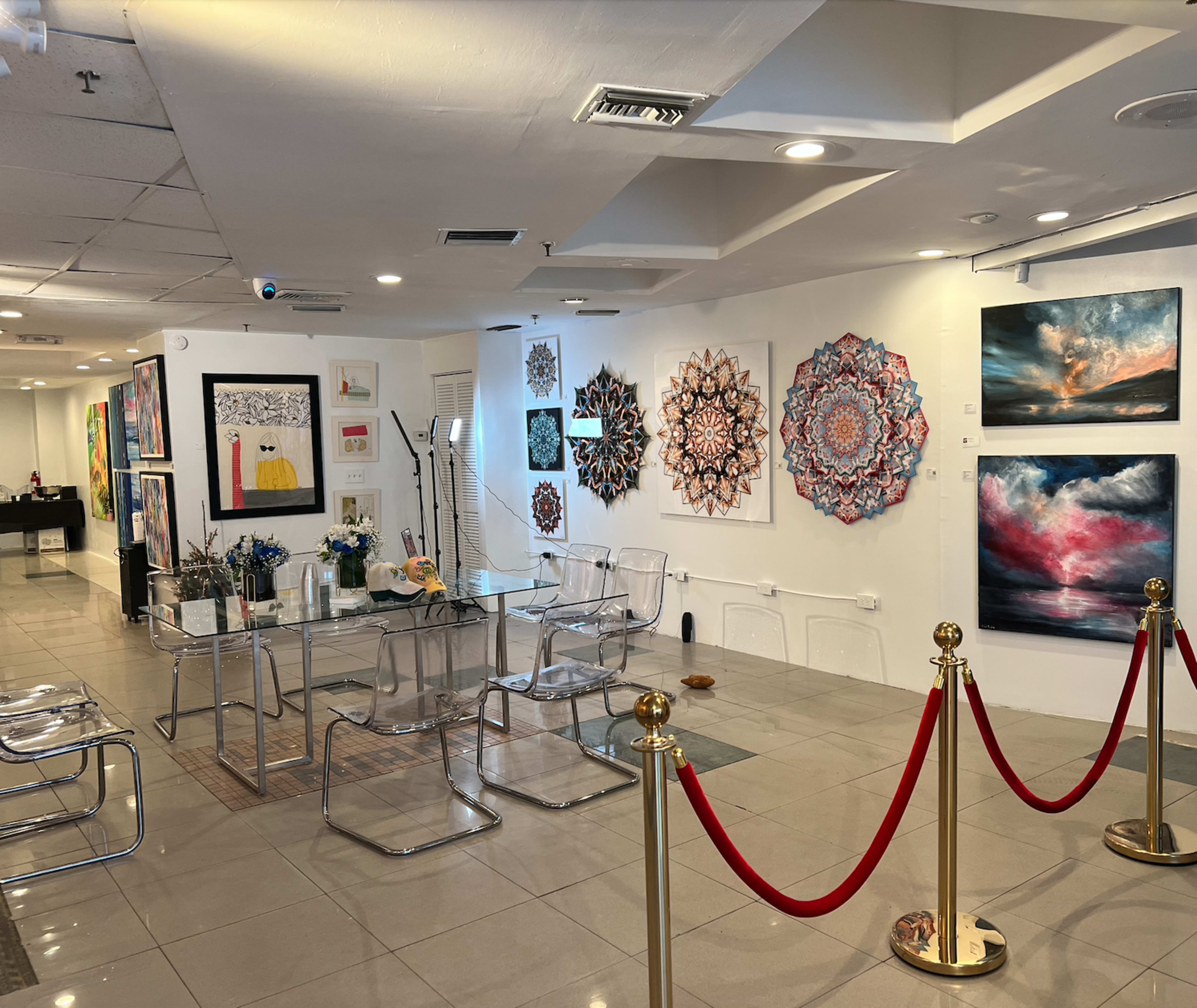 An art gallery featuring various paintings displayed on the walls, with a glass table and chairs in the foreground, and a red rope barrier separating the space.
