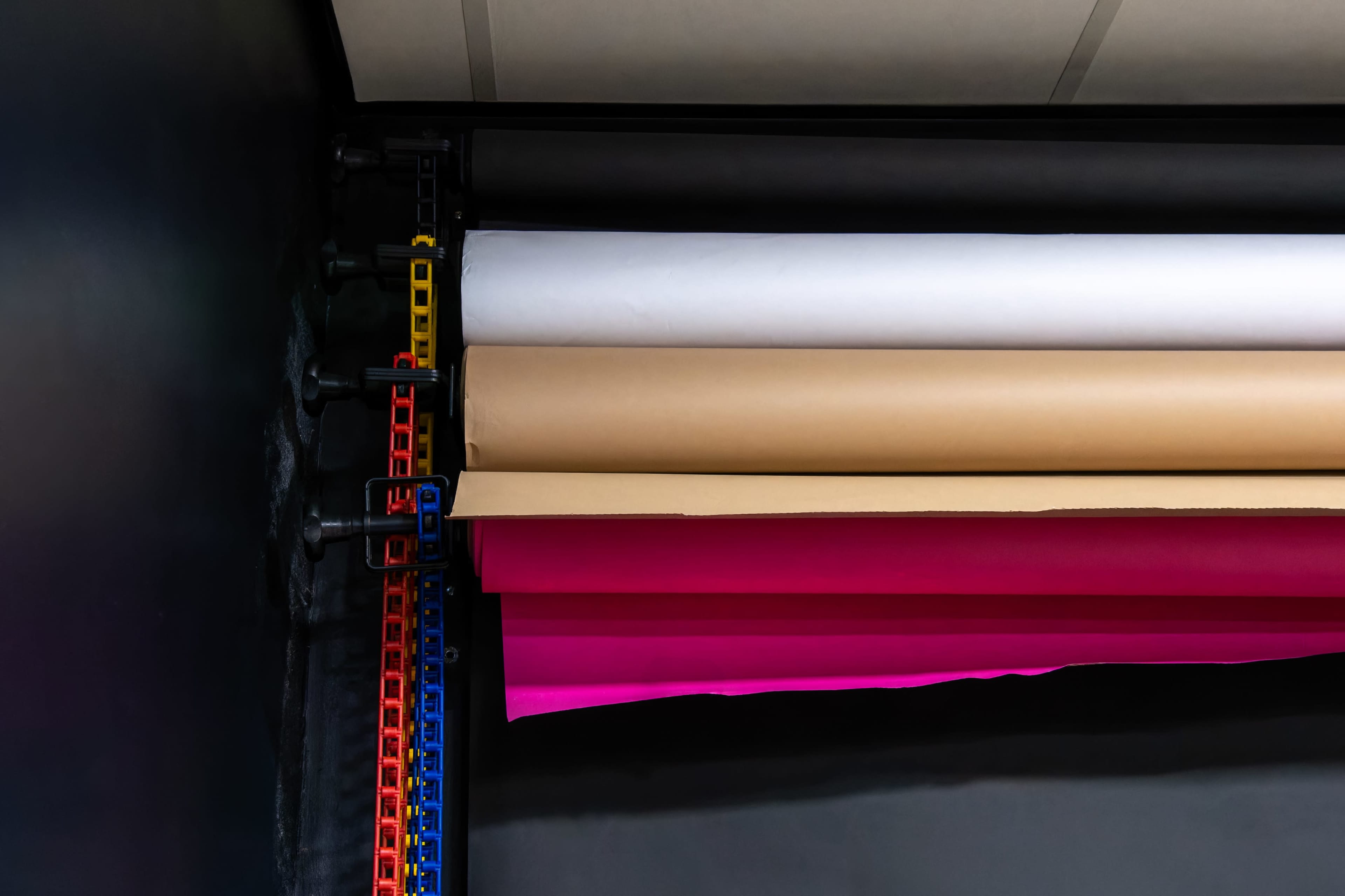 Several rolls of colored paper, including white, brown, and pink, are stored on a rack against a dark wall.