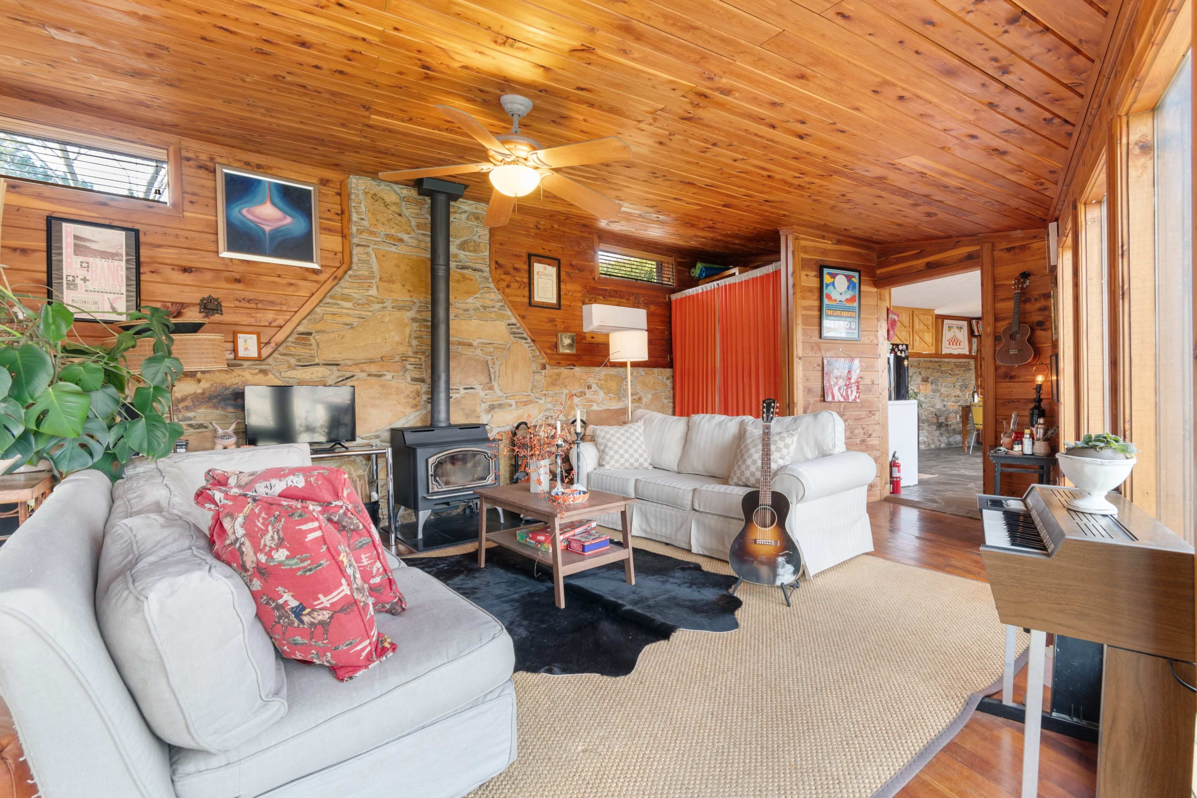 A cozy living room with wooden walls and ceiling, featuring a stone fireplace, two light-colored sofas, and a guitar resting on a black rug near a coffee table.