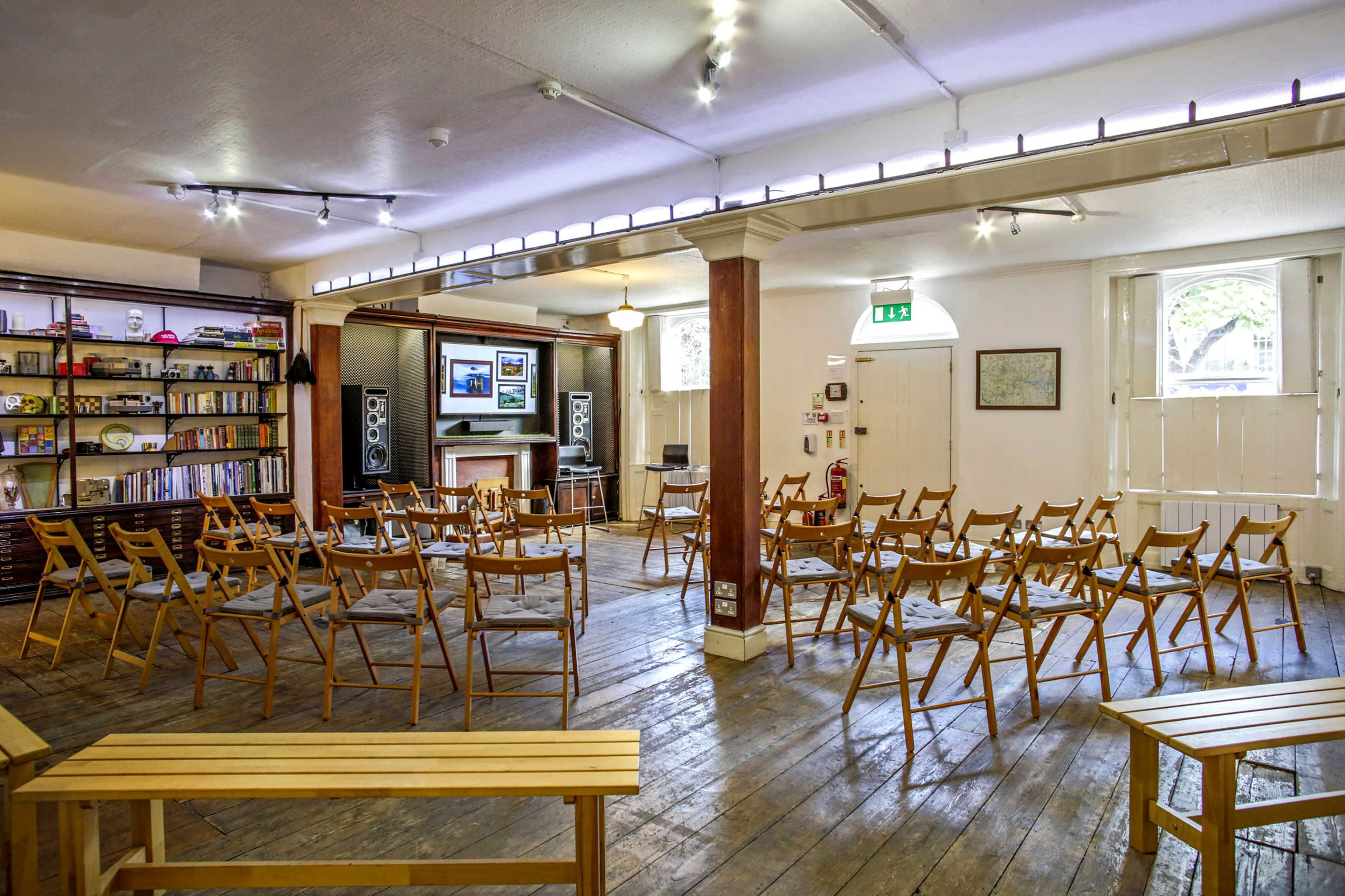 An empty room with wooden flooring arranged with rows of folding chairs facing a stage area and a television mounted on the wall.