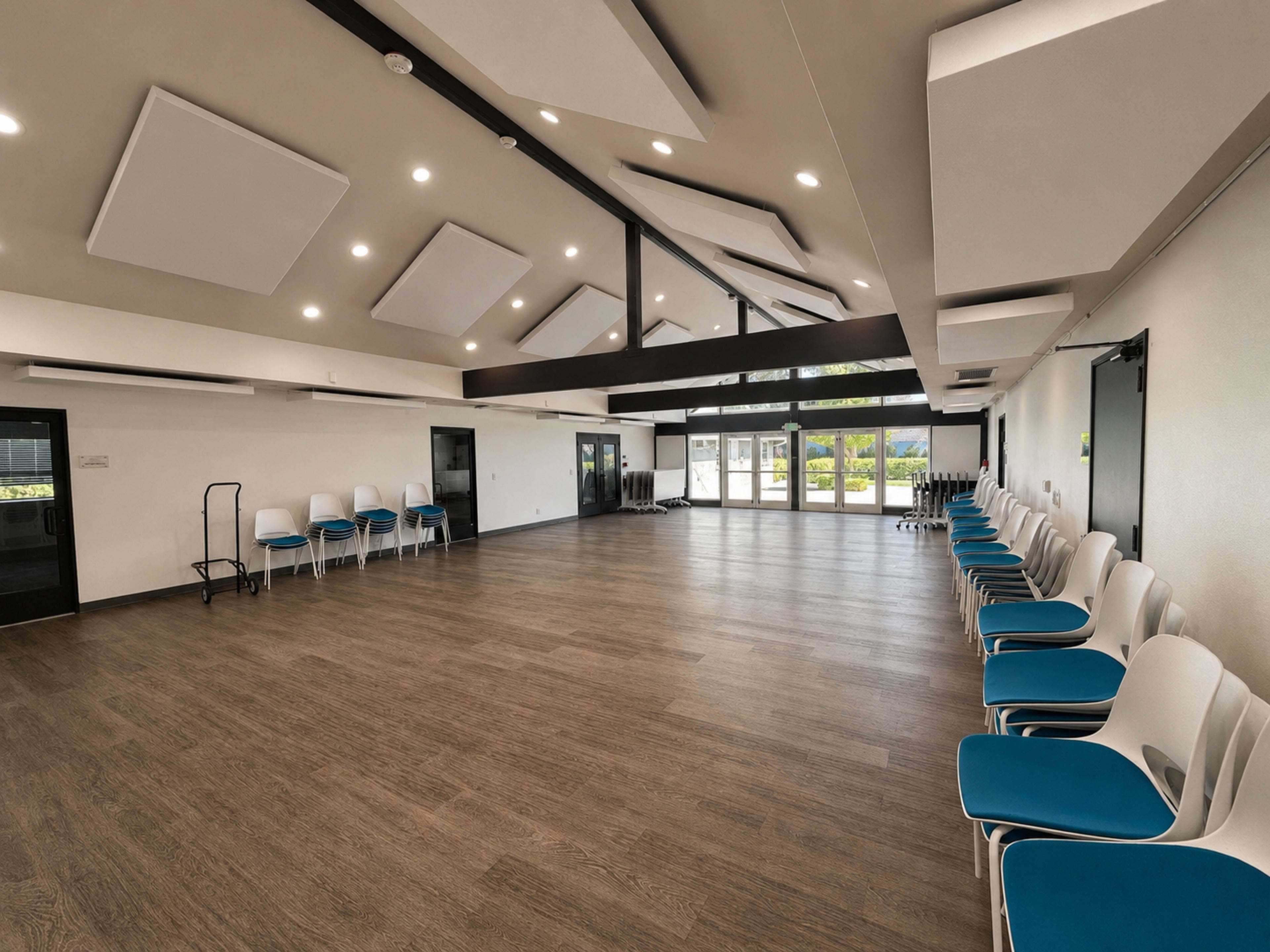 The image shows an empty community room with rows of white chairs along the walls and a high ceiling featuring modern acoustic panels.