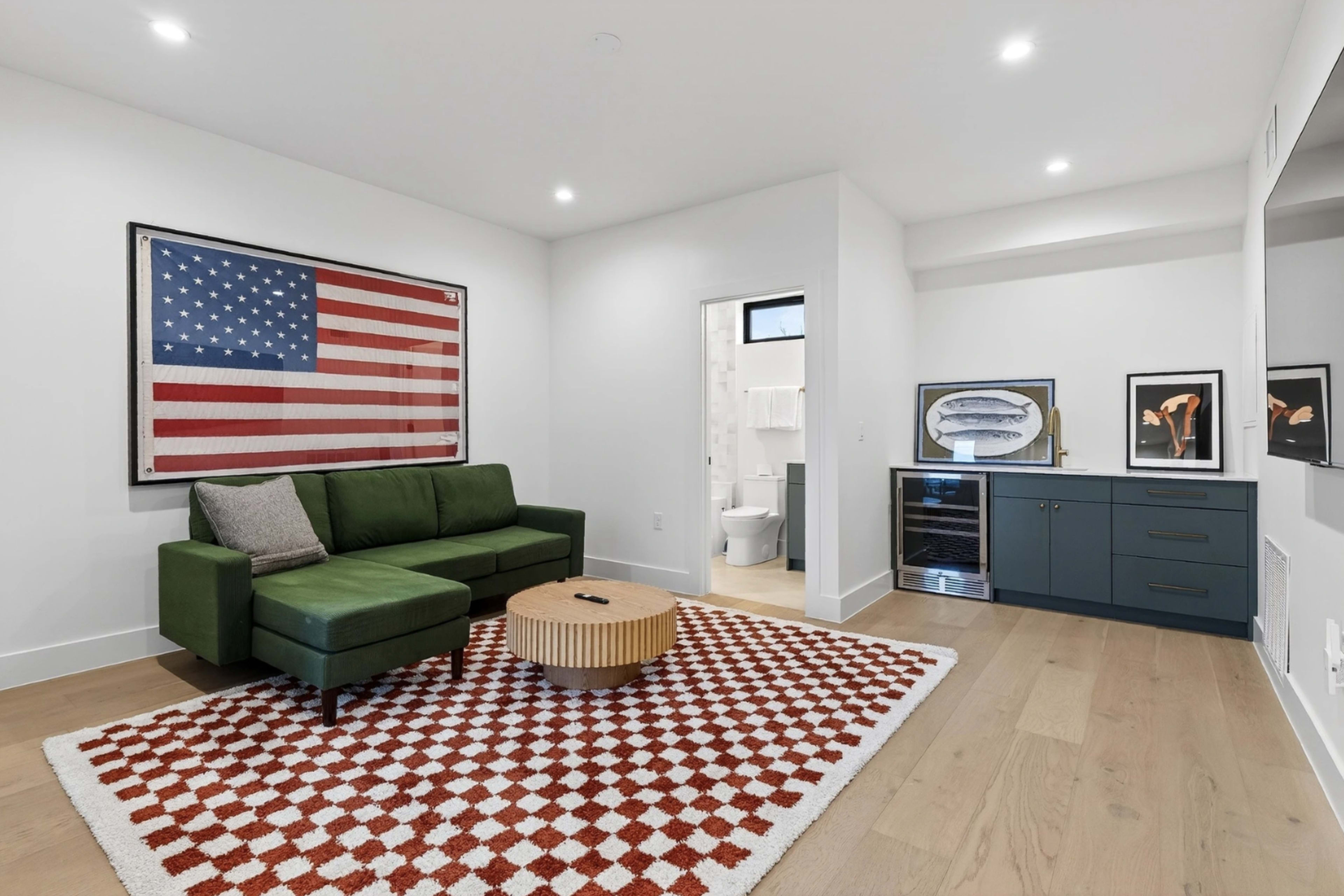 The image shows a modern living space featuring a green sectional couch, a round wooden coffee table, an American flag artwork on the wall, and a small kitchenette area with a sink and refrigerator.